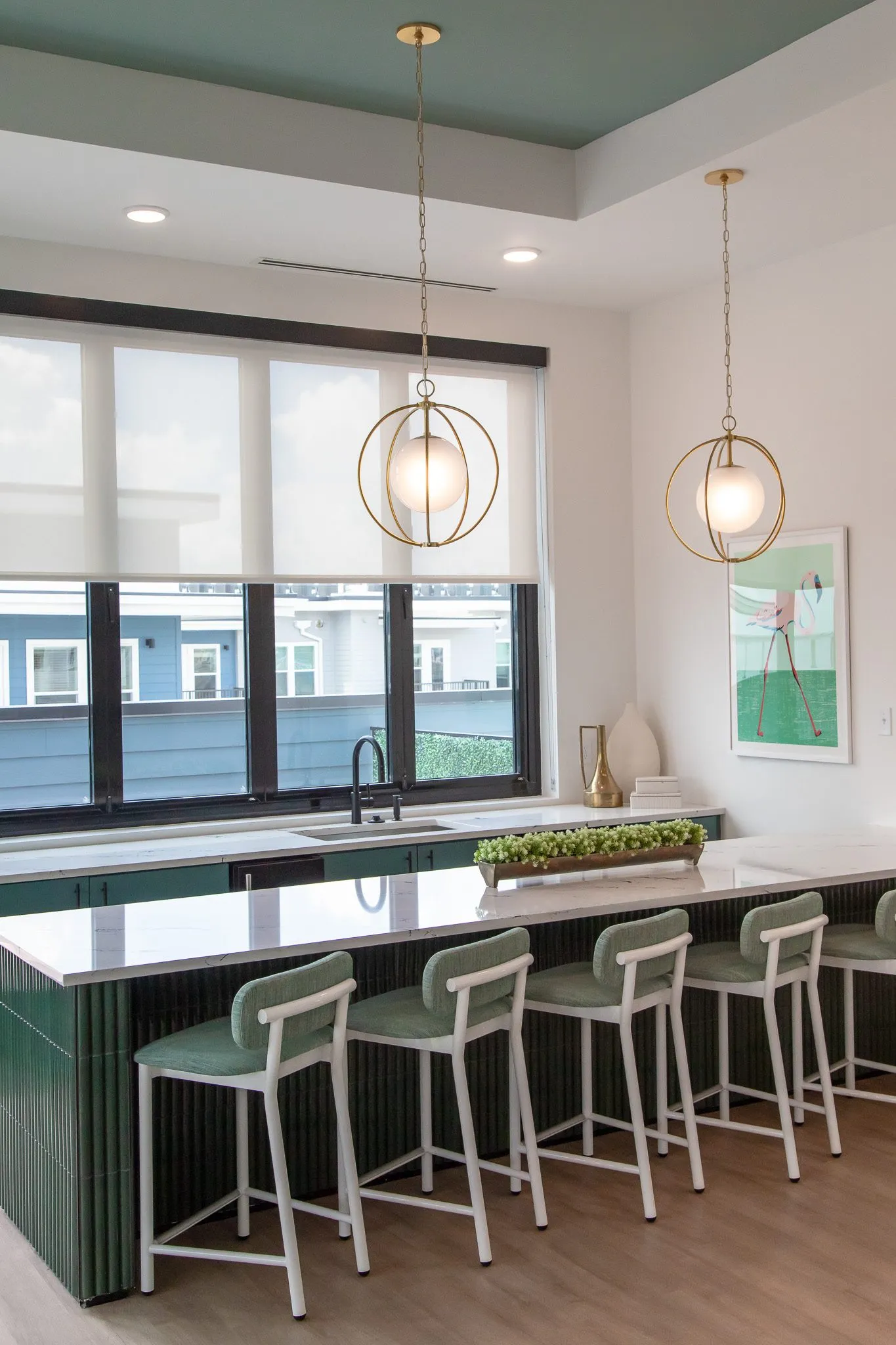 Multifamily kitchen island seen from the seating side, green stools under brass orb pendants before a wide window