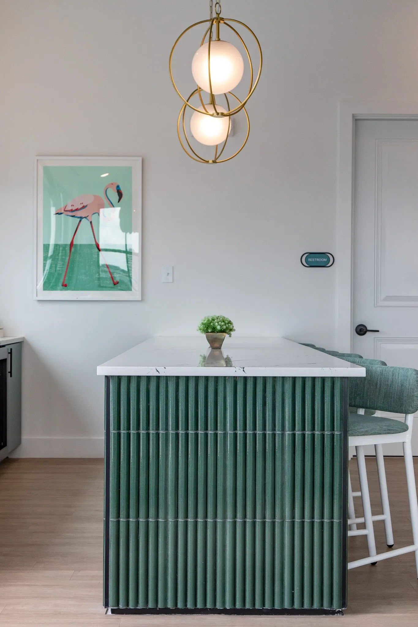 Multifamily interior detail: fluted green tile island end below brass orb pendants and a flamingo print