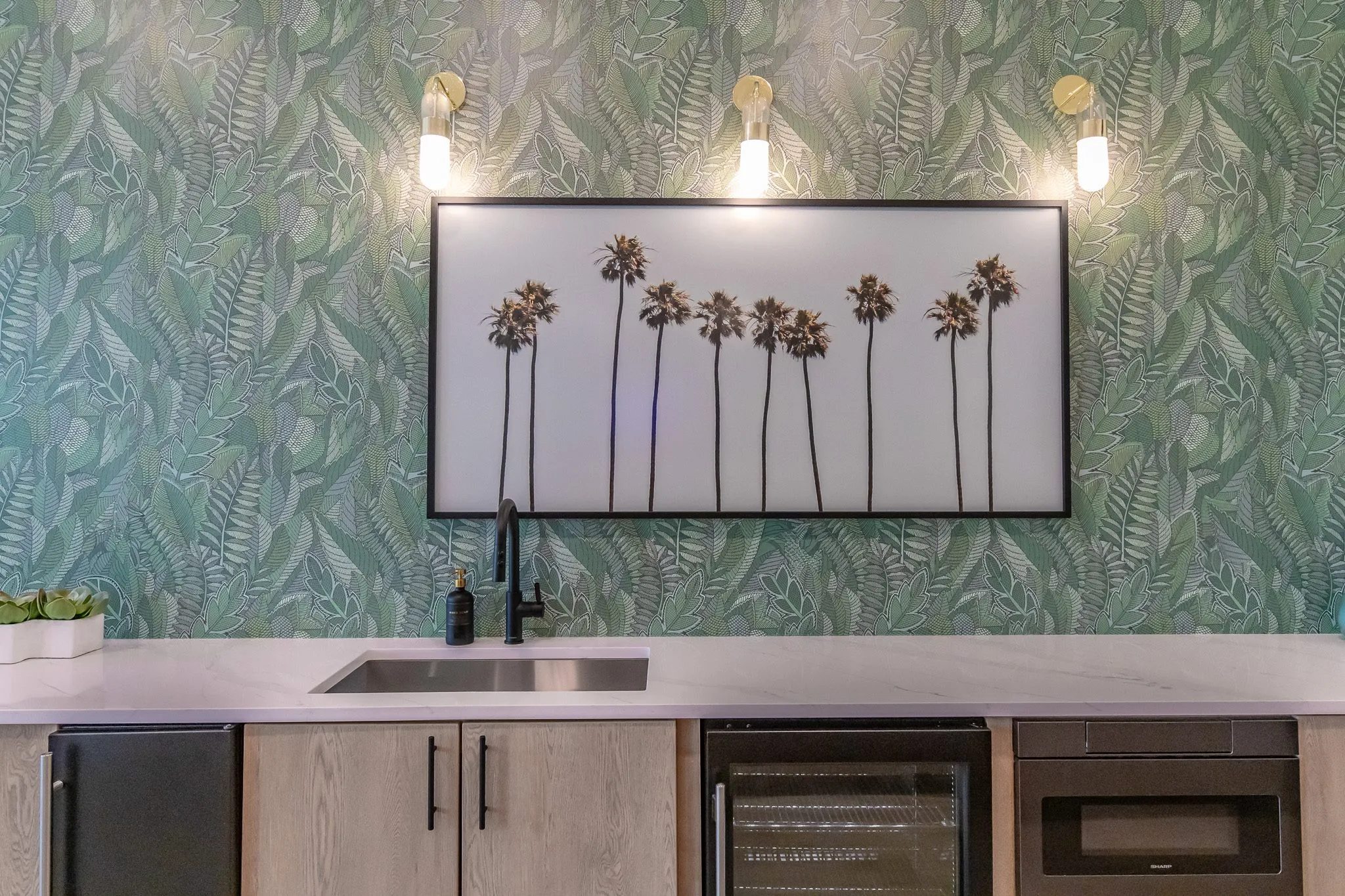 Multifamily amenity kitchenette with a stainless sink and wine fridge below a palm print and brass sconces on leaf wallpaper