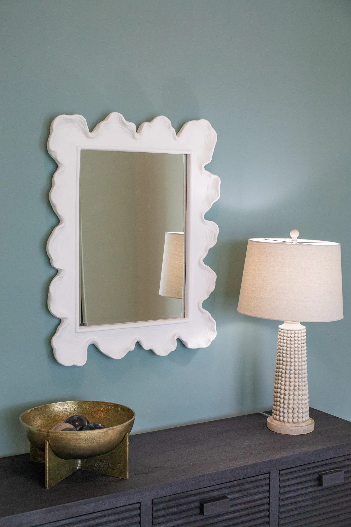 Multifamily interior detail: scalloped white mirror and a beaded lamp on a dark dresser against a blue grey wall
