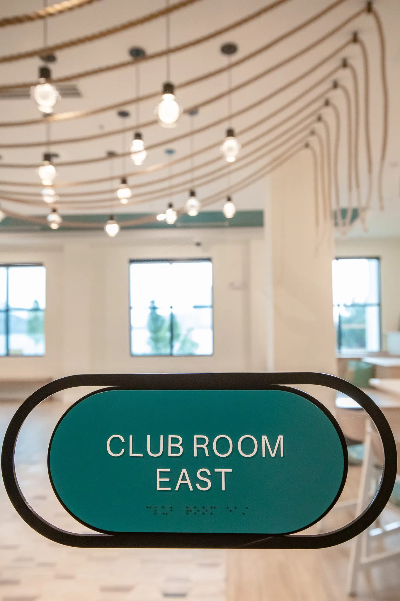 Multifamily amenity door sign reading Club Room East with draped rope pendants and windows beyond