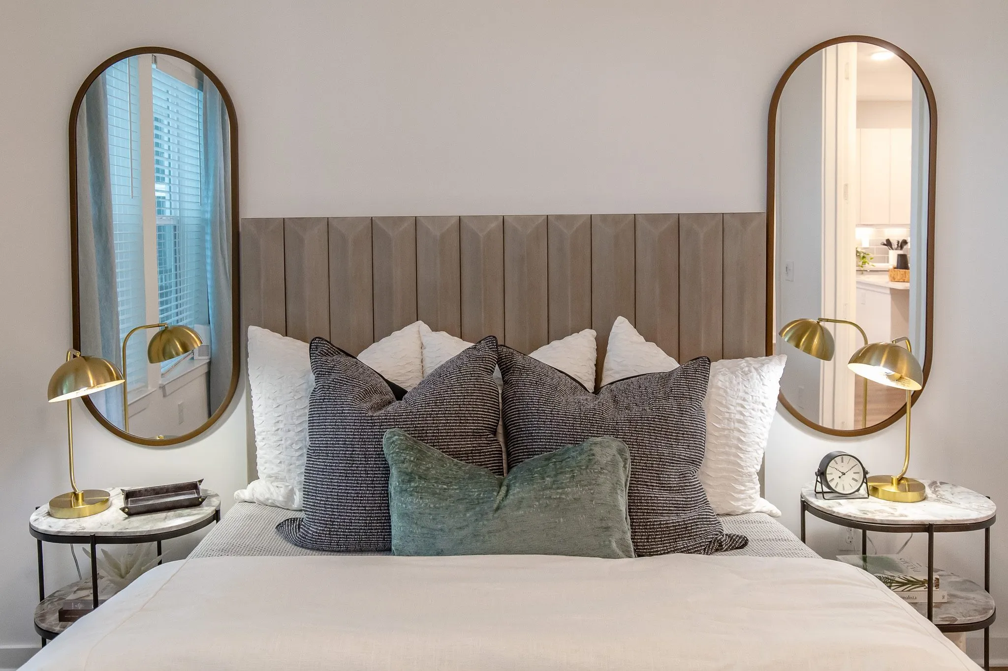 Multifamily model apartment bedroom with a channelled wood headboard between oval mirrors, brass lamps on marble nightstands