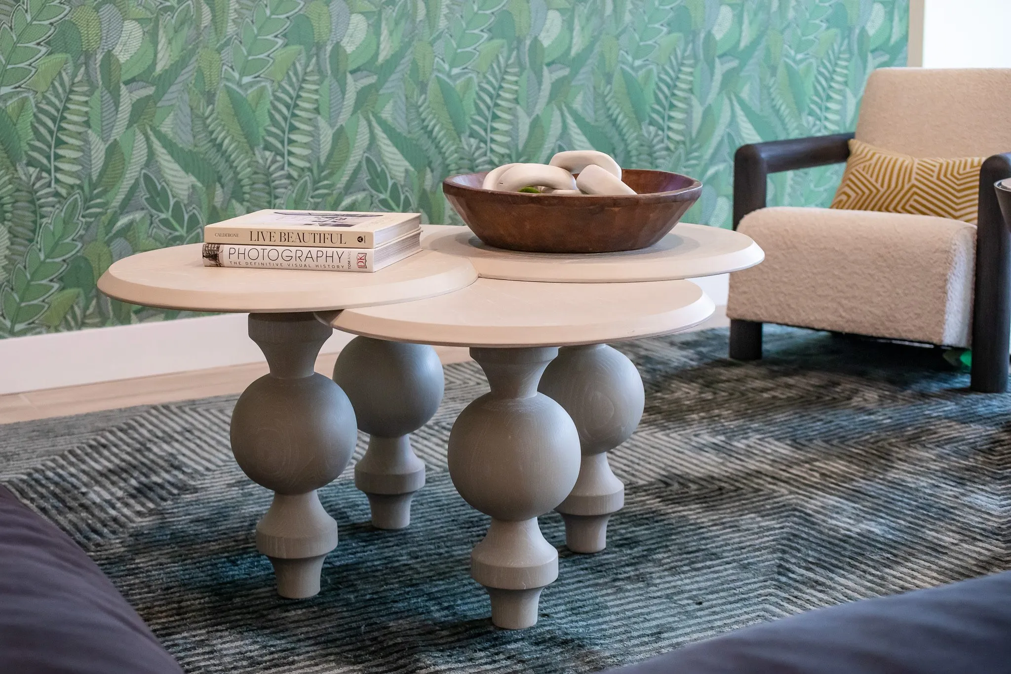 Multifamily interior detail: grey nesting coffee tables with a wood bowl and books before leaf wallpaper and a boucle chair