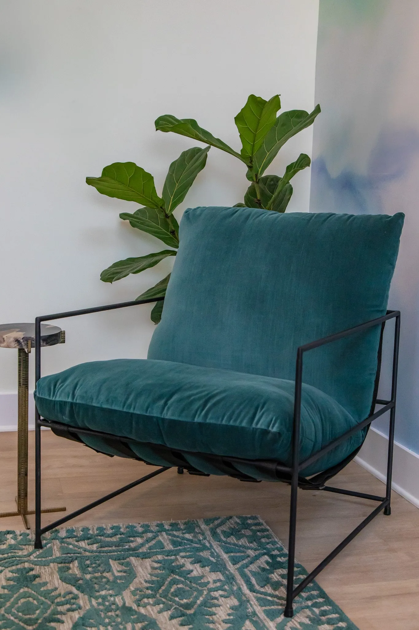 Multifamily interior detail: teal velvet sling armchair on a black metal frame with a fiddle leaf fig and patterned rug