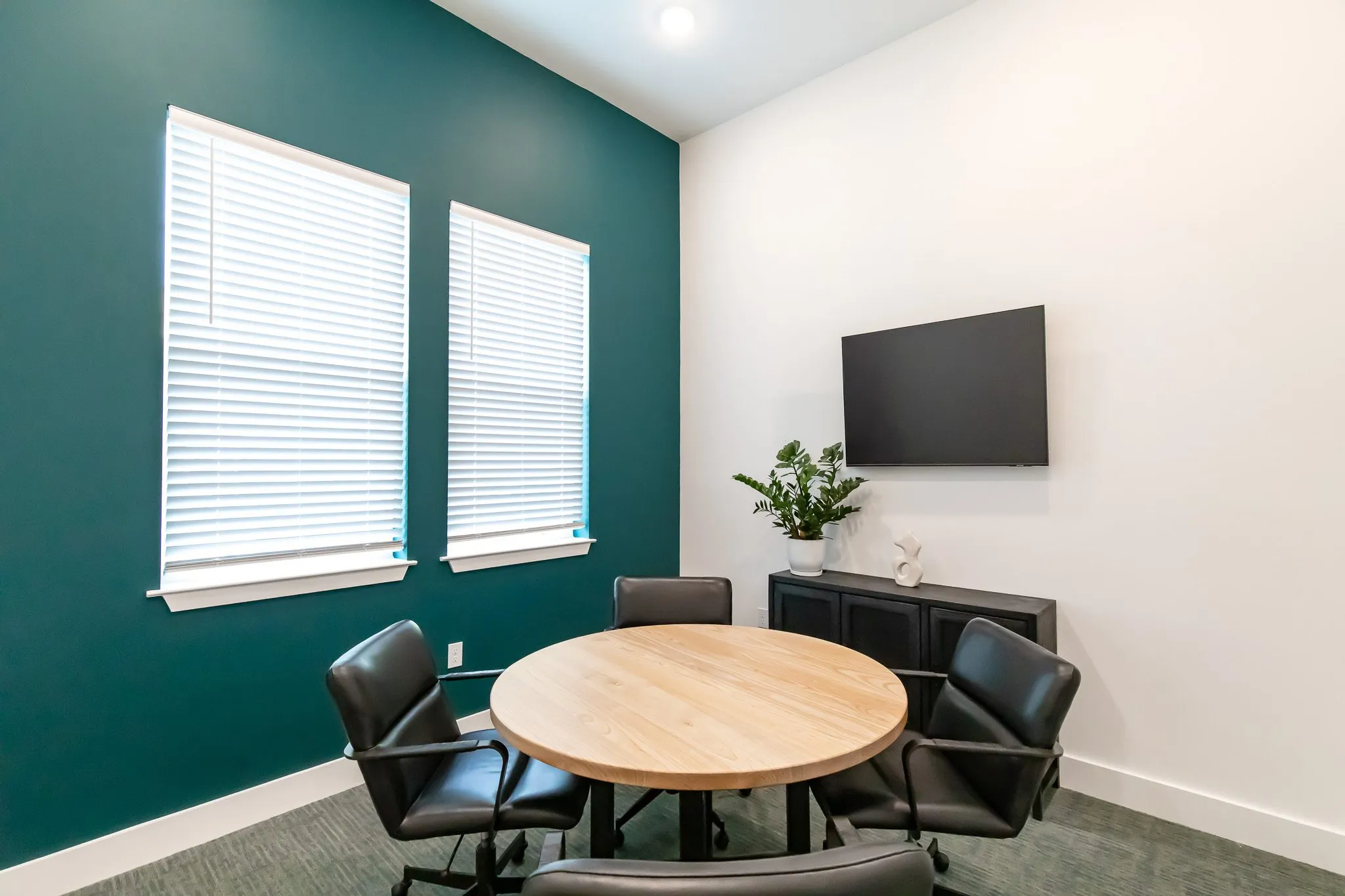 Multifamily amenity meeting room with a round wood table, black leather chairs, a teal accent wall and a wall mounted TV
