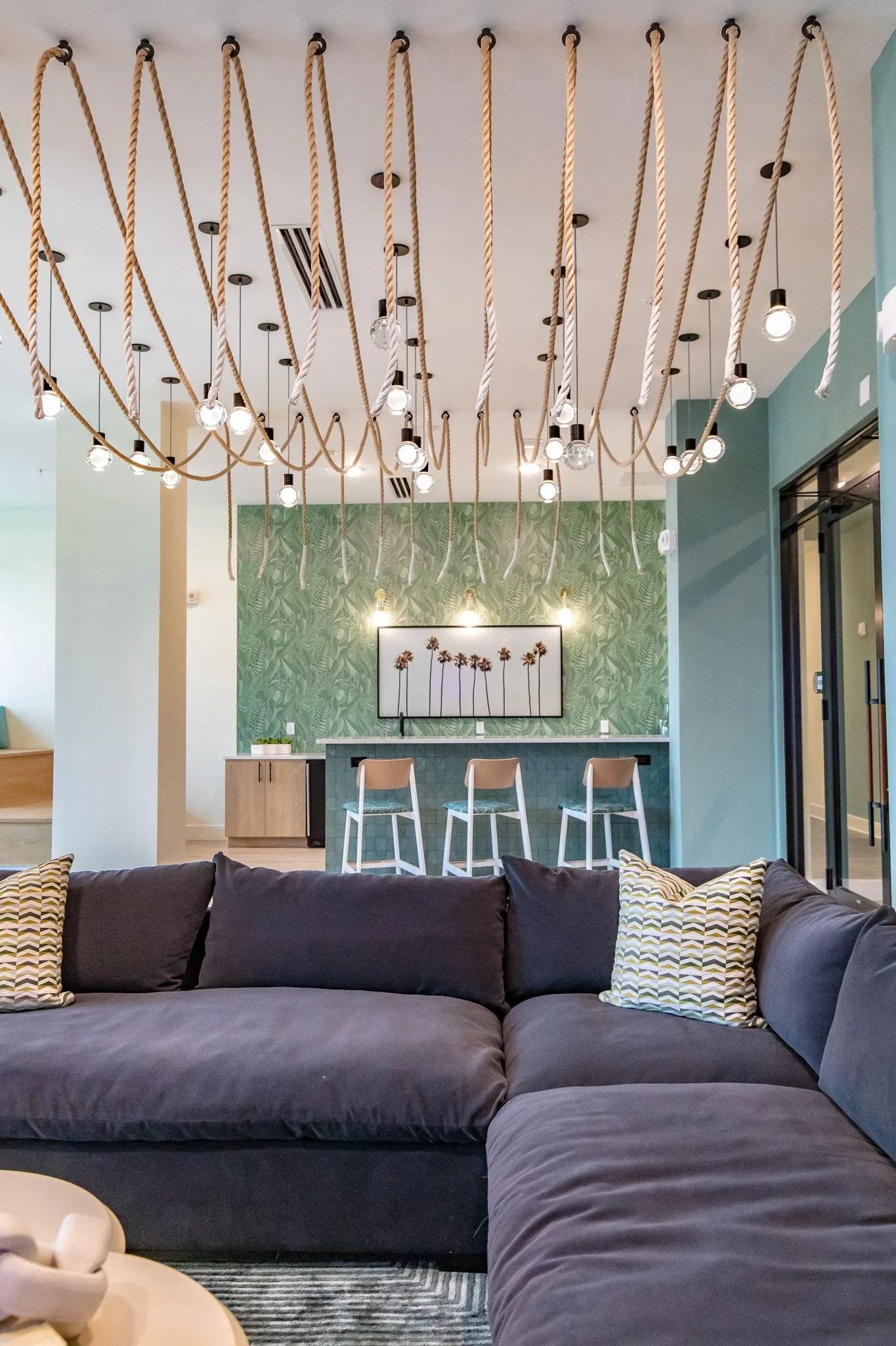 Multifamily amenity lounge with a navy sectional under draped rope and bulb pendants, a bar with stools on leaf wallpaper