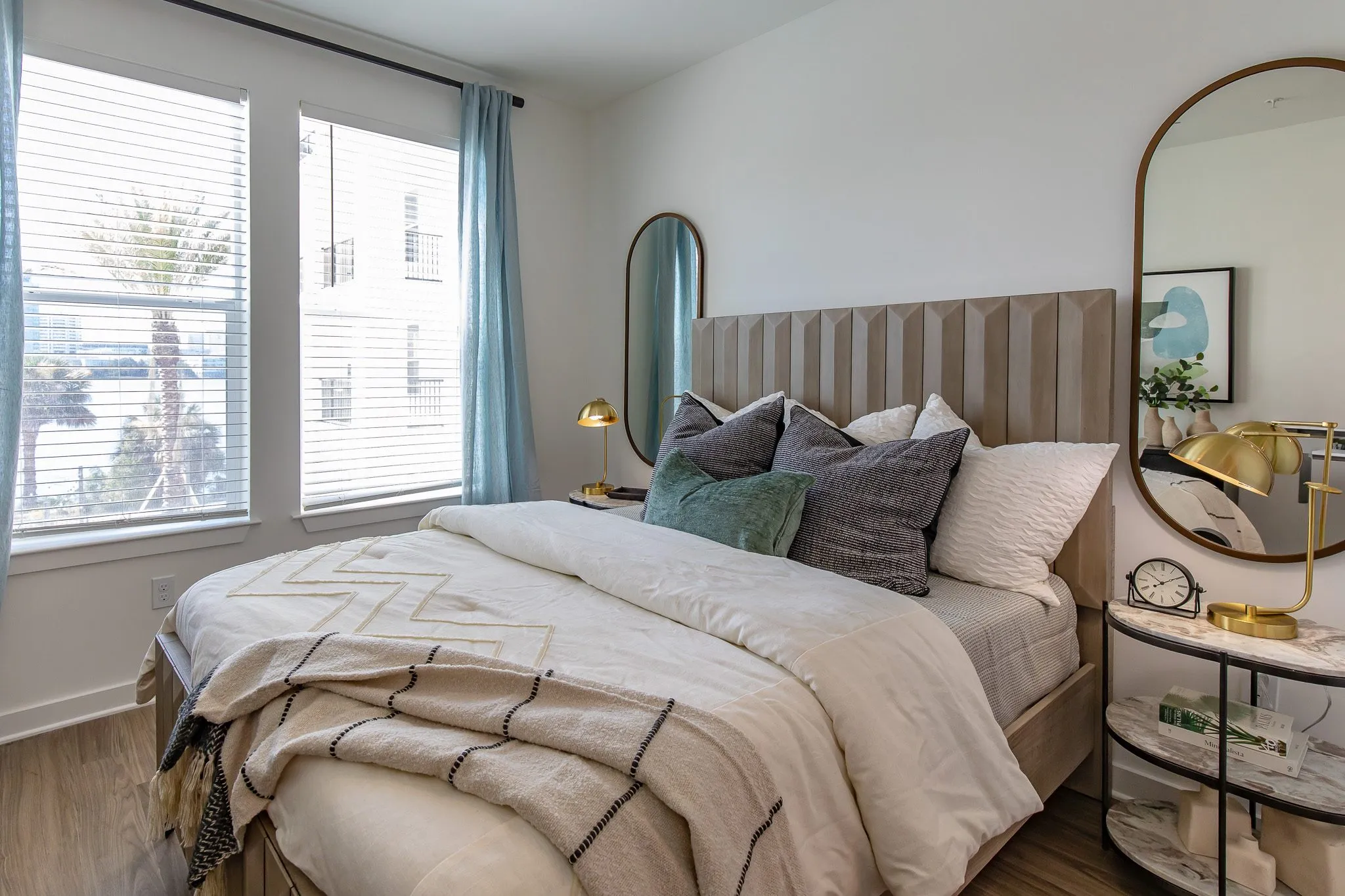 Multifamily model apartment bedroom with a chevron throw on the bed, curtained windows and an oval mirror above a lamp