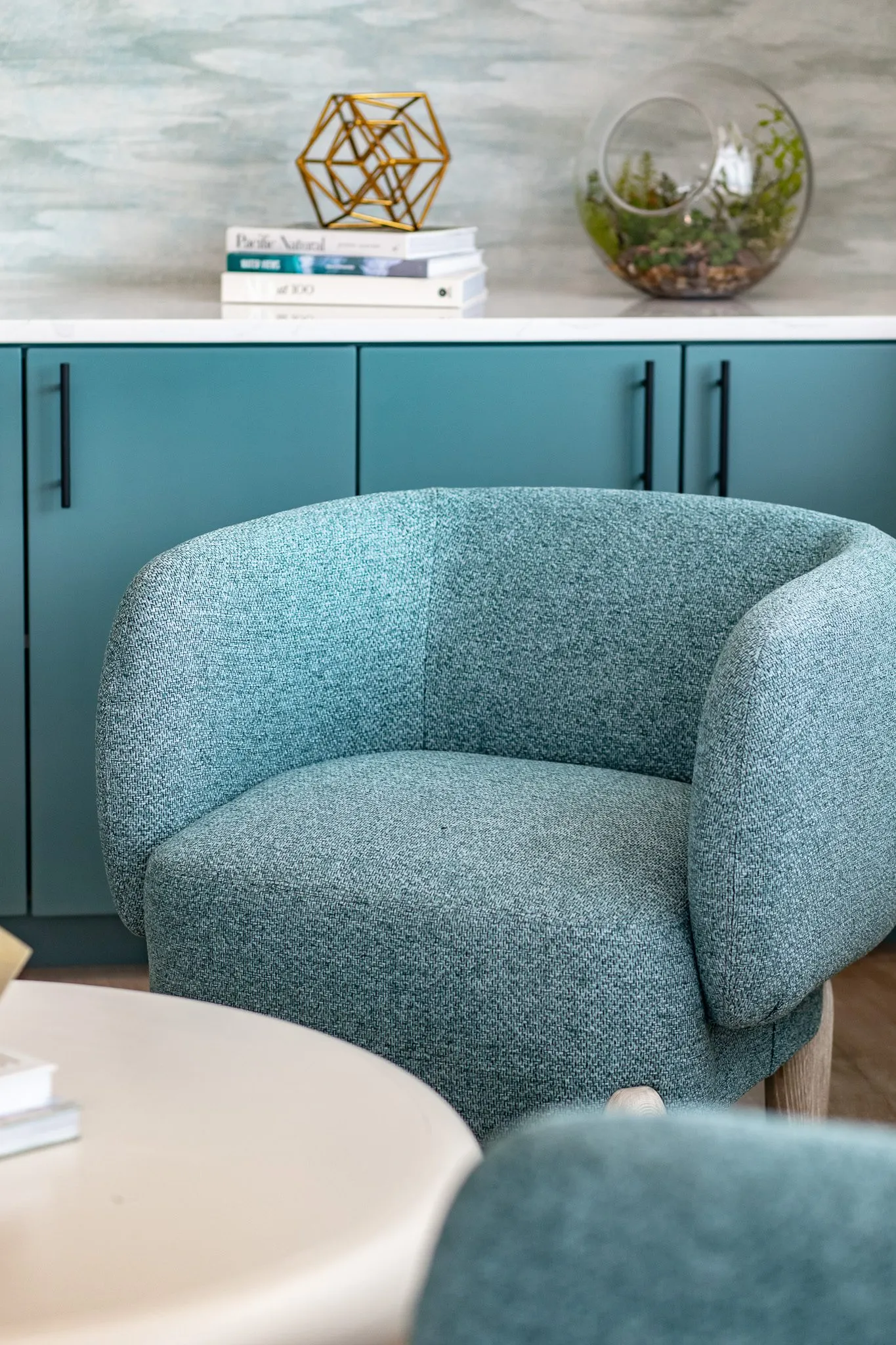 Multifamily interior detail: teal boucle barrel chair before teal cabinets with books, a brass sculpture and a terrarium