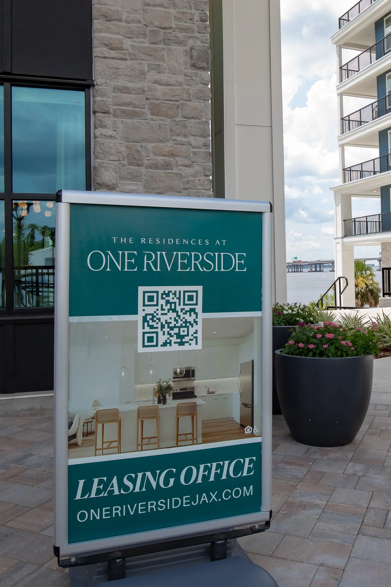 Multifamily community exterior: teal A-frame leasing office sign with a QR code on the paved entry beside the building