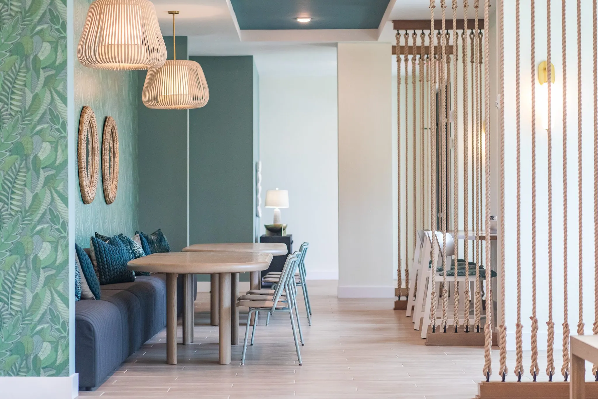 Multifamily amenity banquette hall from beside the rope screen, navy seating and oval tables under slatted pendants