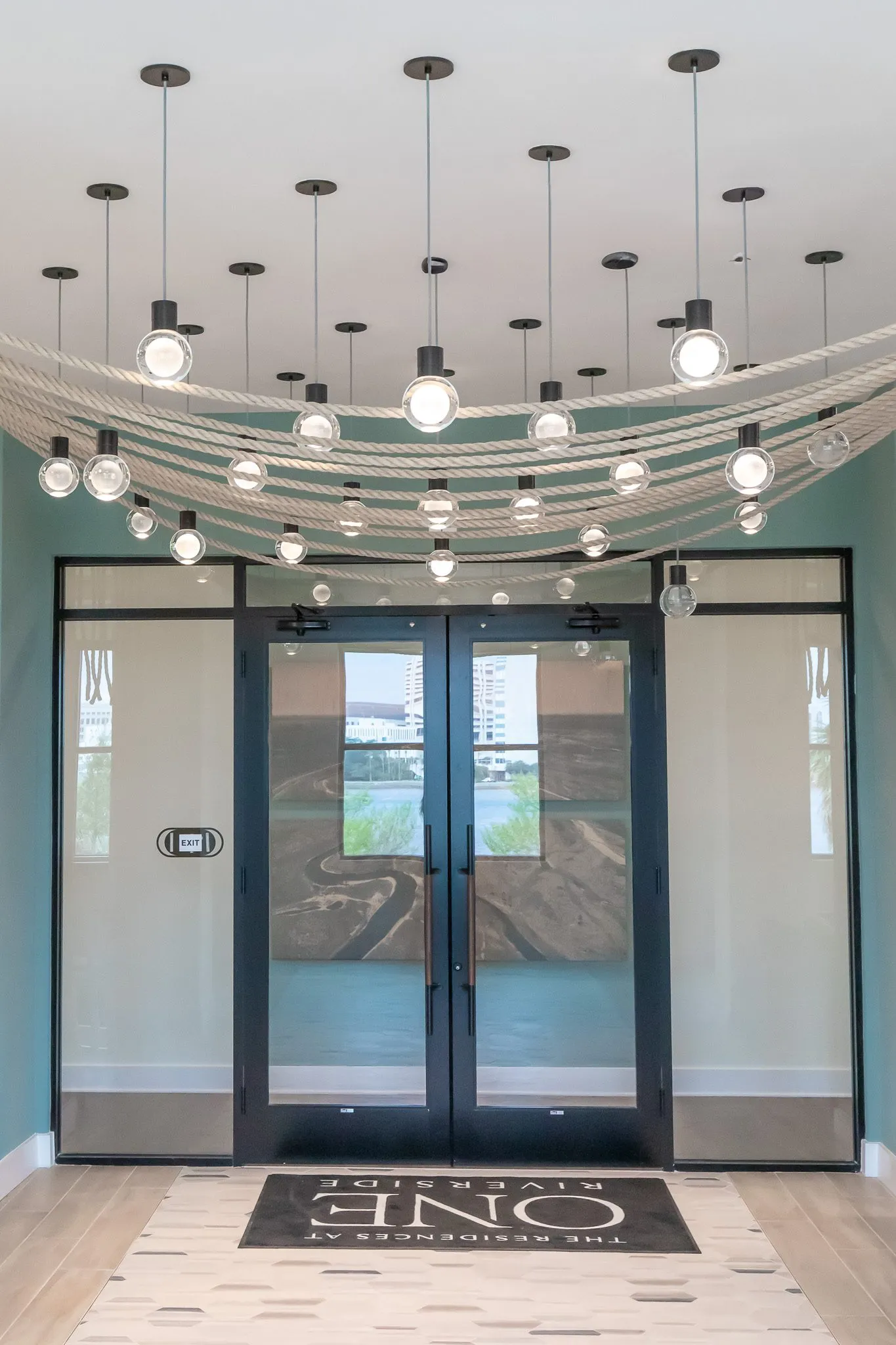 Multifamily amenity entry vestibule with glass double doors, a logo mat and a canopy of draped rope with bulb pendants