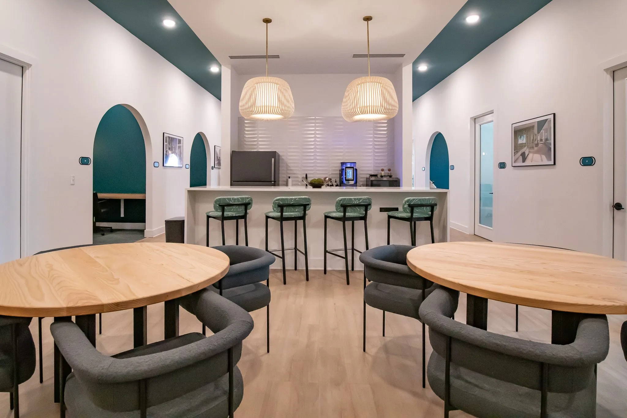 Multifamily amenity break room with round wood tables and grey barrel chairs, green stools at a counter below pendants