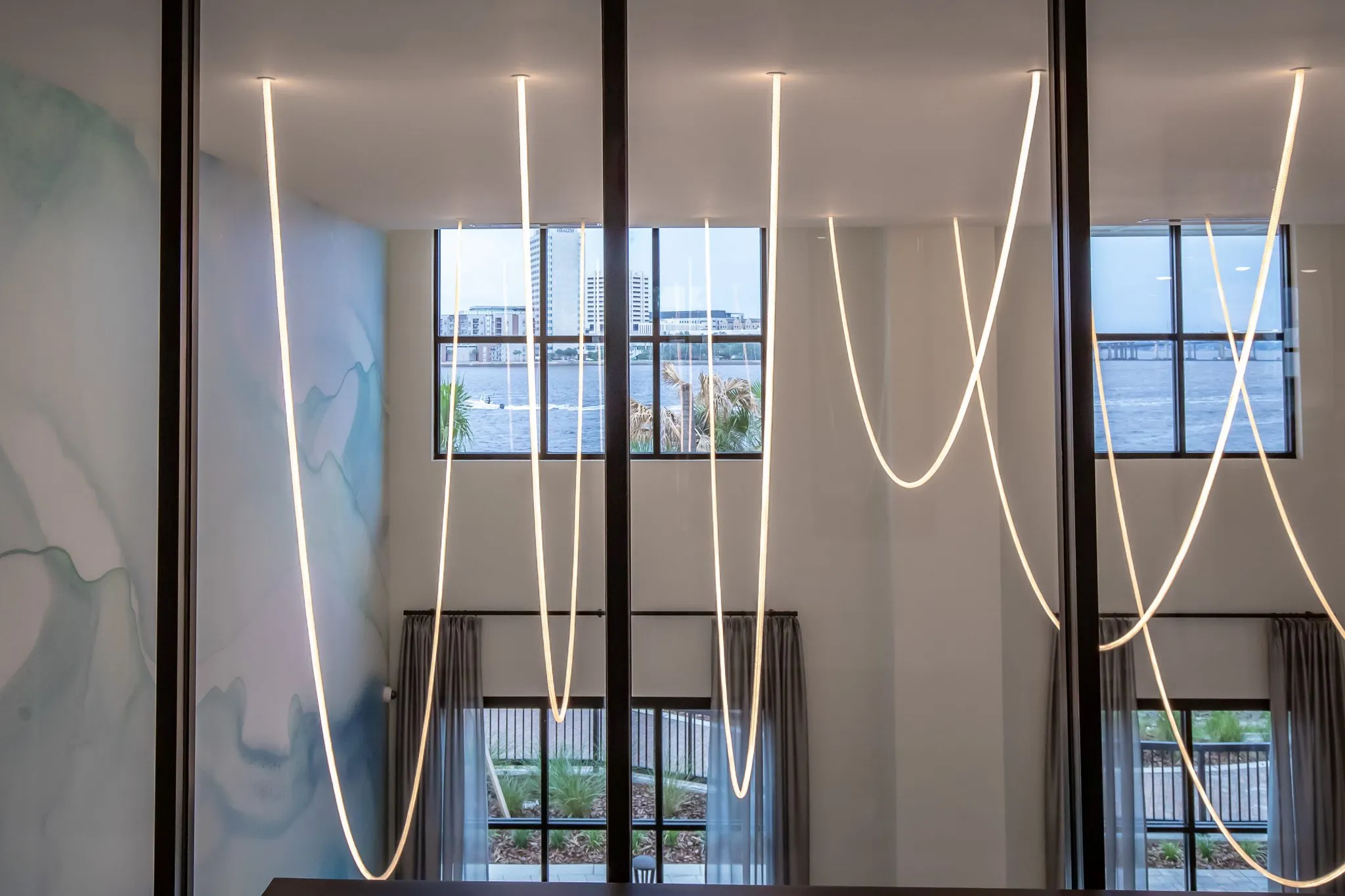 Multifamily interior: looping linear light strands hanging past tall windows with a river view, a watercolor mural at left