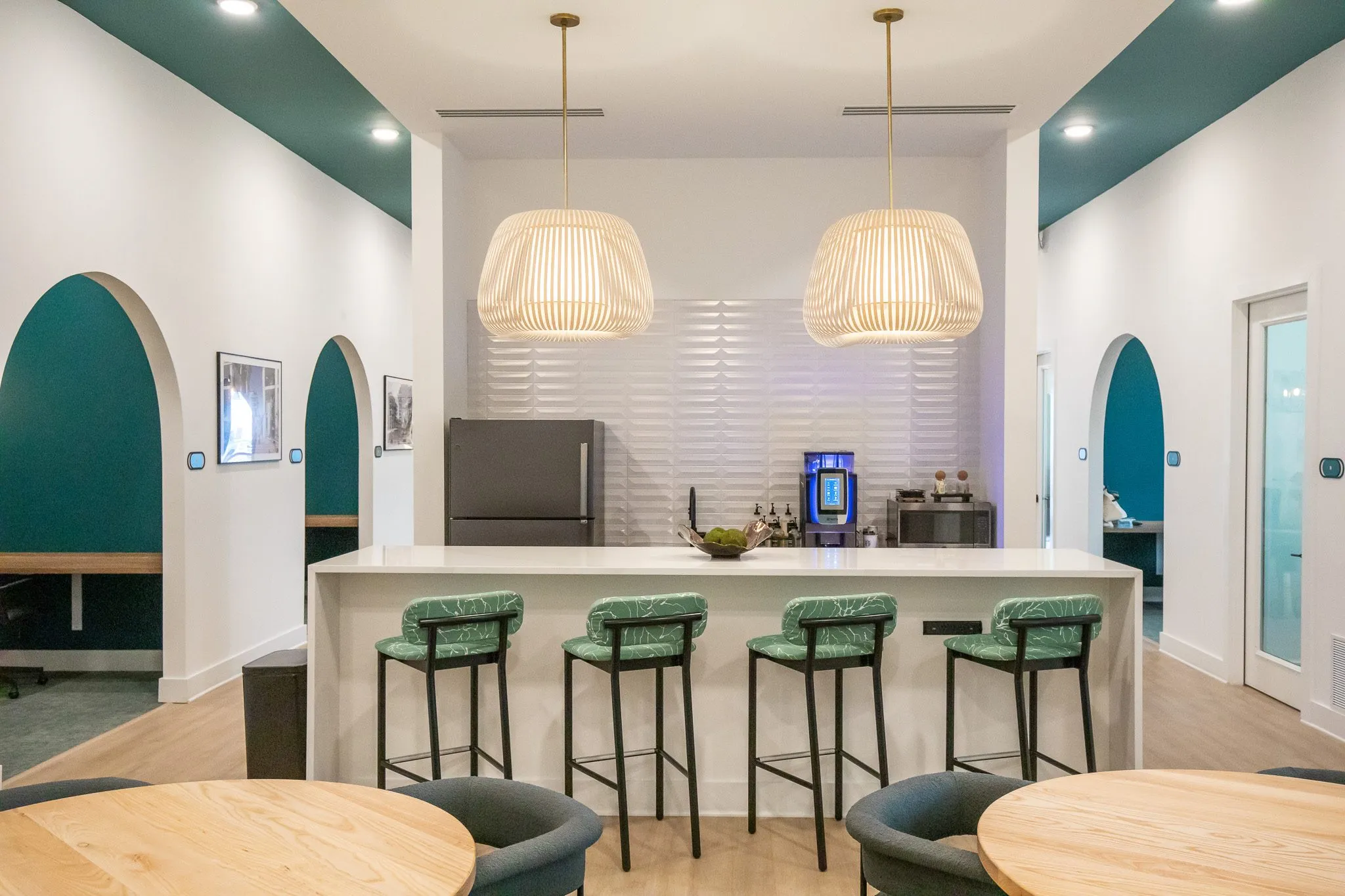 Multifamily amenity break room with a white island and leaf print stools under slatted pendants, teal arched doorways