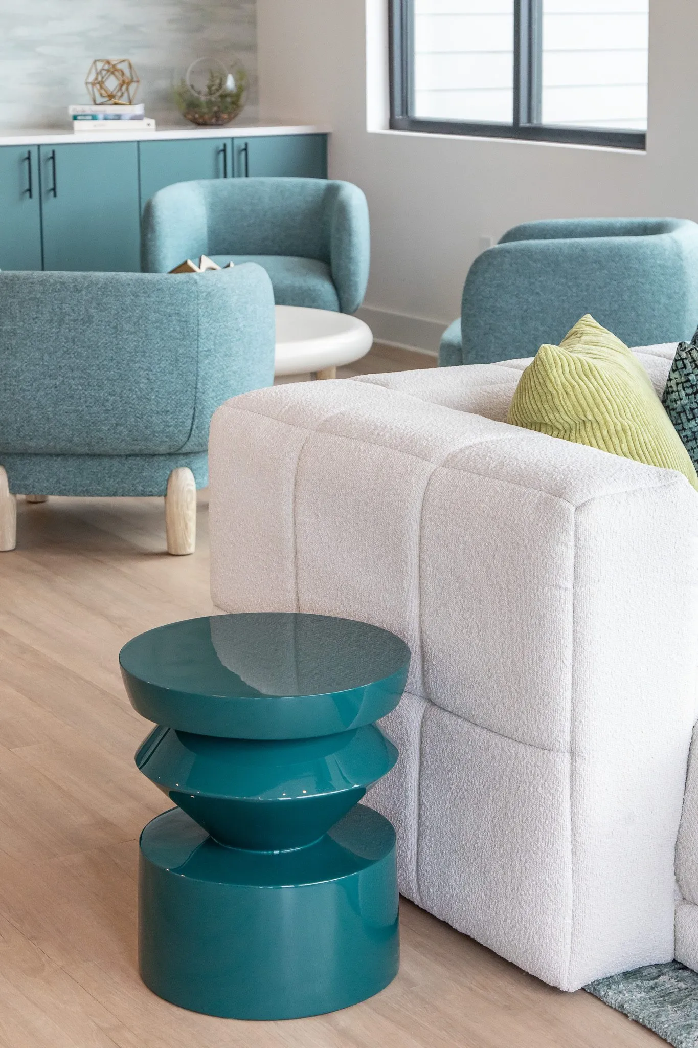 Multifamily interior detail: teal lacquered side table by a white boucle sectional, teal barrel chairs and cabinets beyond