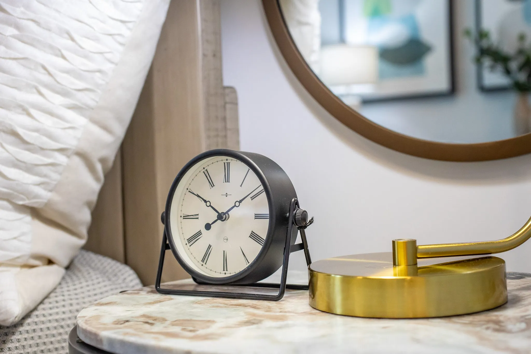 Multifamily apartment bedroom detail: black alarm clock and brass lamp on a marble nightstand below a round mirror
