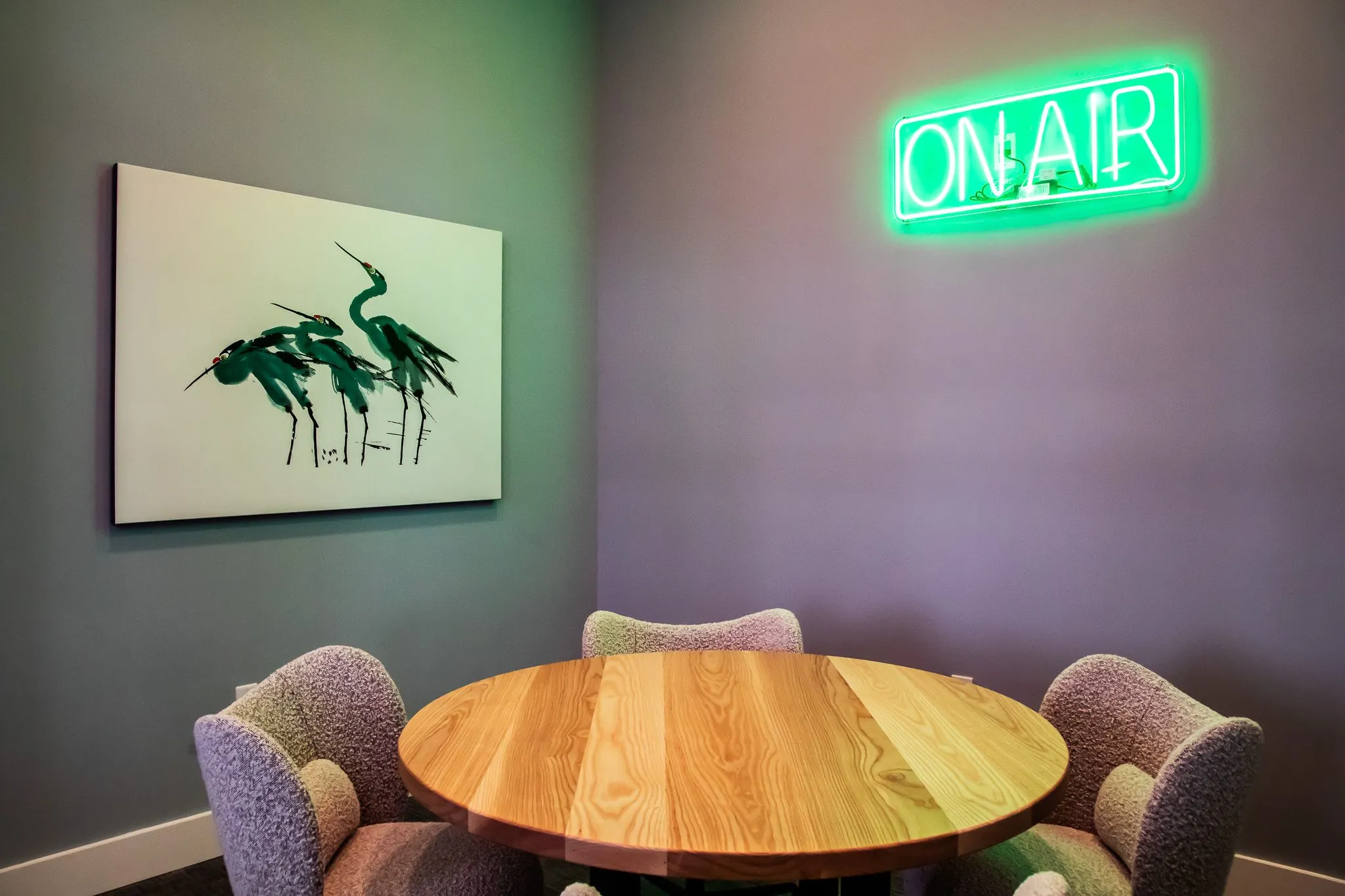 Multifamily amenity podcast room with lilac boucle chairs at a round wood table, a heron print and a green neon On Air sign