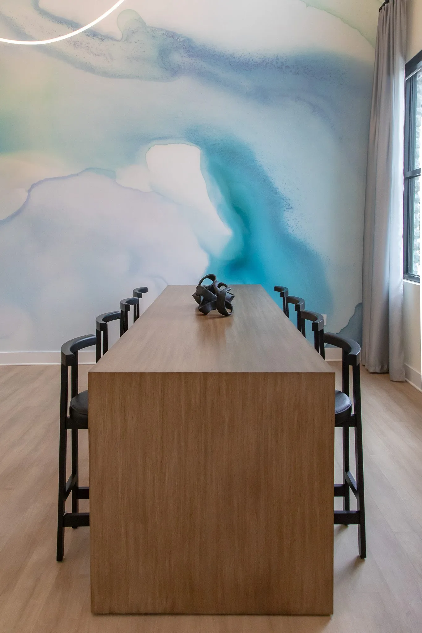 Multifamily amenity lounge with a long wood communal table and black stools before a blue watercolor mural