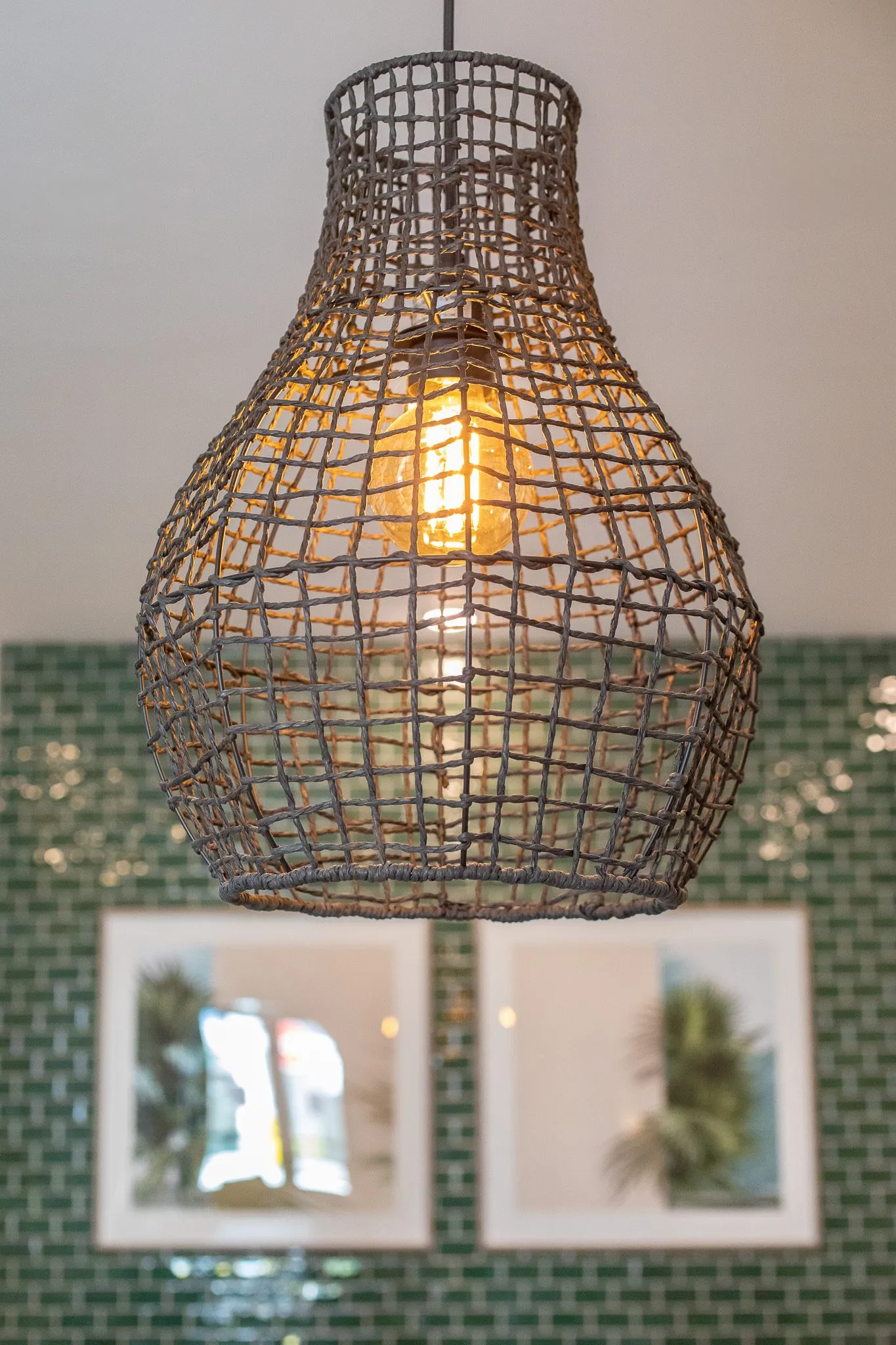 Multifamily interior detail: woven wire pendant with a bulb before green tile and framed palm prints