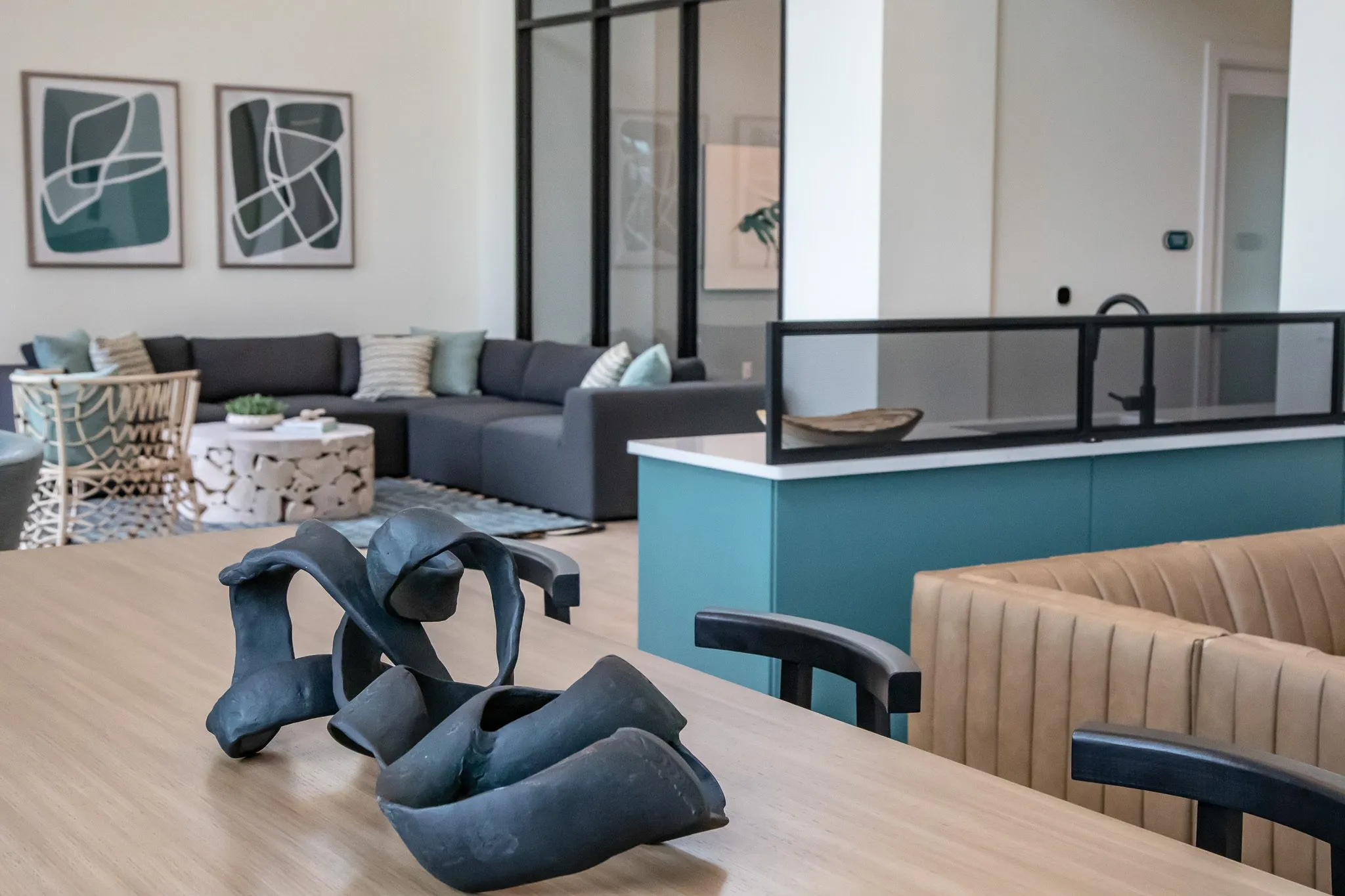 Multifamily interior detail: black ribbon sculpture on a wood table, tan banquette and grey sectional with prints beyond