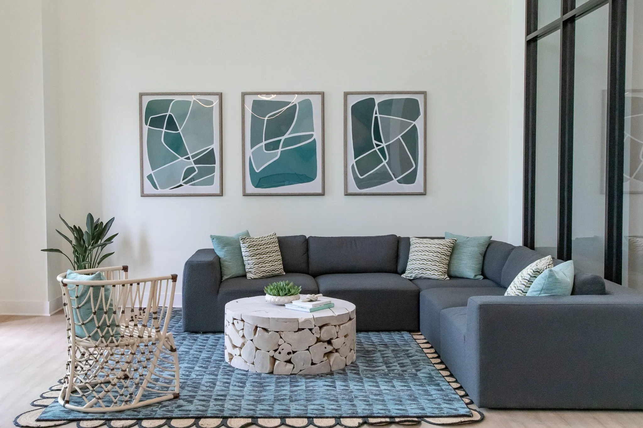 Multifamily amenity lounge with a grey sectional, driftwood drum table and rattan chair below three teal abstract prints