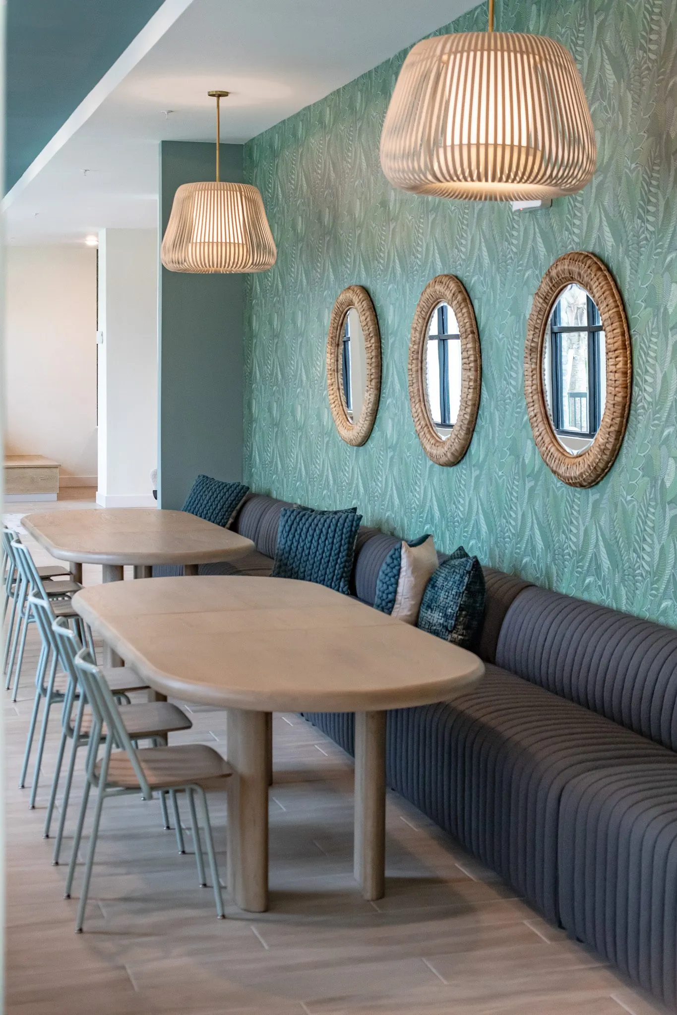 Multifamily amenity banquette hall with navy seating, oval wood tables and three round rattan mirrors on leaf wallpaper