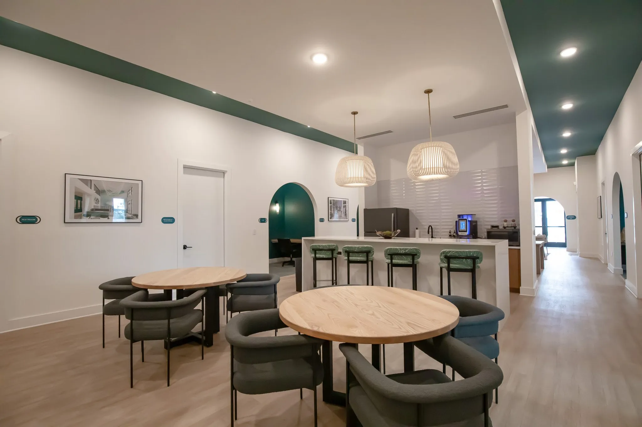 Multifamily amenity break room from the corridor, wood tables with grey chairs, green stools at a counter under pendants