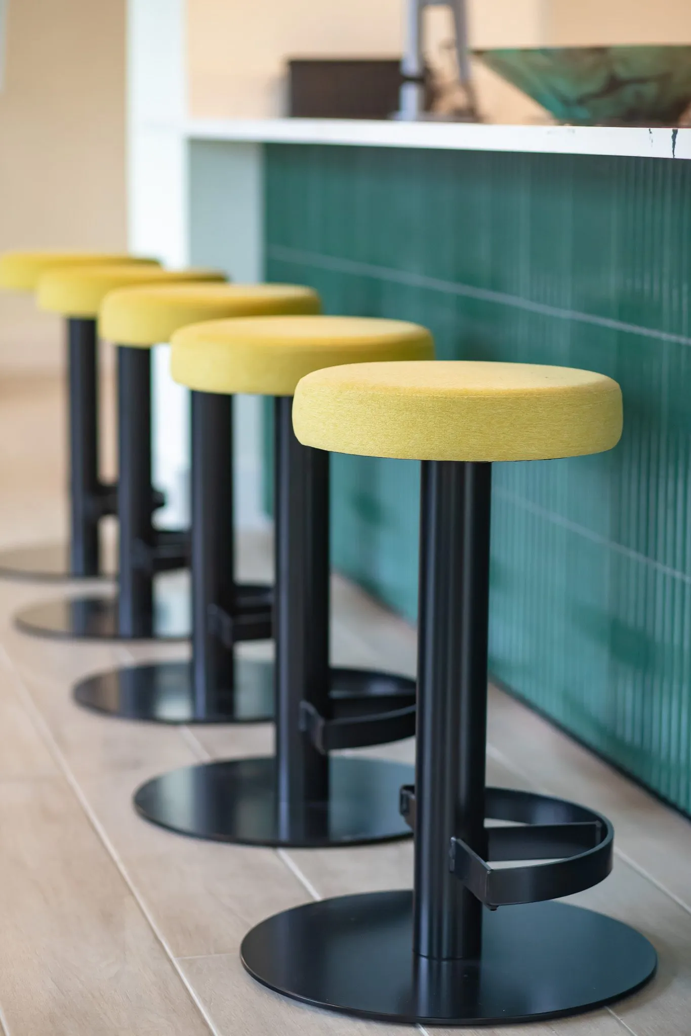 Multifamily interior detail: low angle along a row of yellow topped stools on black pedestals beside fluted green tile