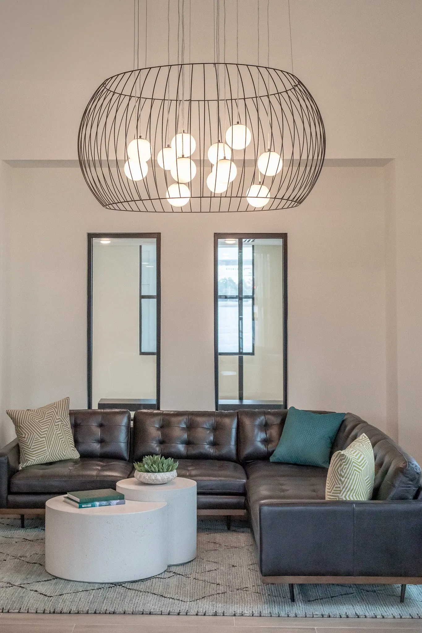 Multifamily amenity lounge with a black leather tufted sectional and white drum tables under a wire cage chandelier