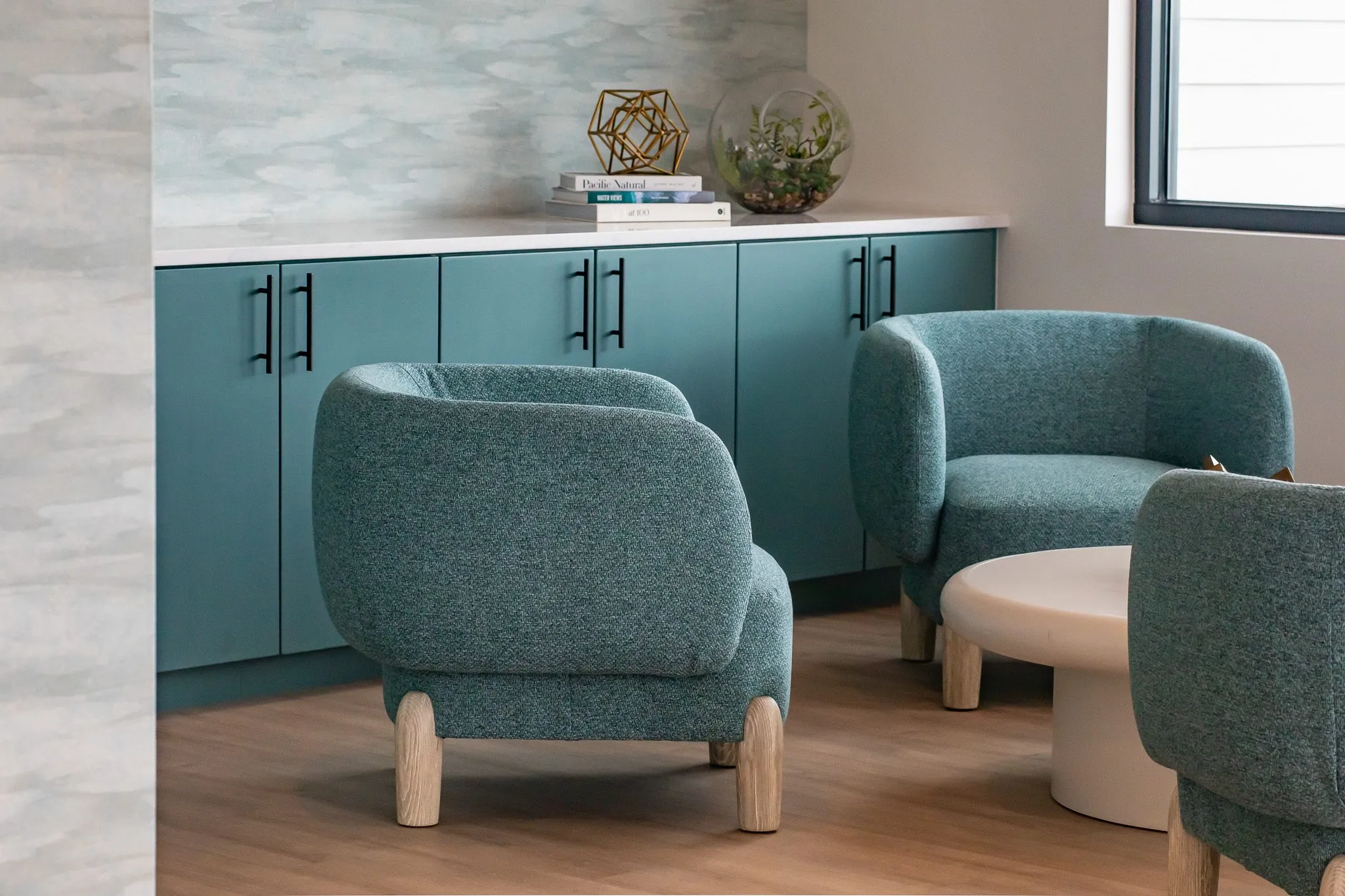 Multifamily interior detail: teal boucle barrel chairs on pale wood legs around a white drum table, teal cabinets beyond