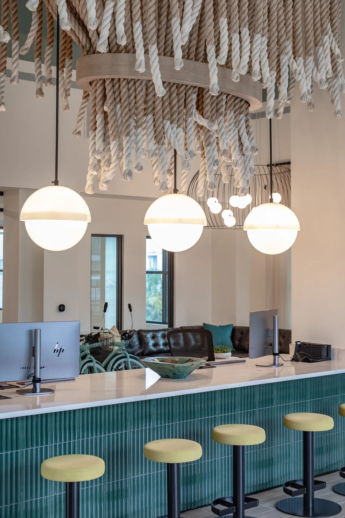 Multifamily leasing desk with monitors on a white counter over fluted teal tile, yellow stools and globe pendants