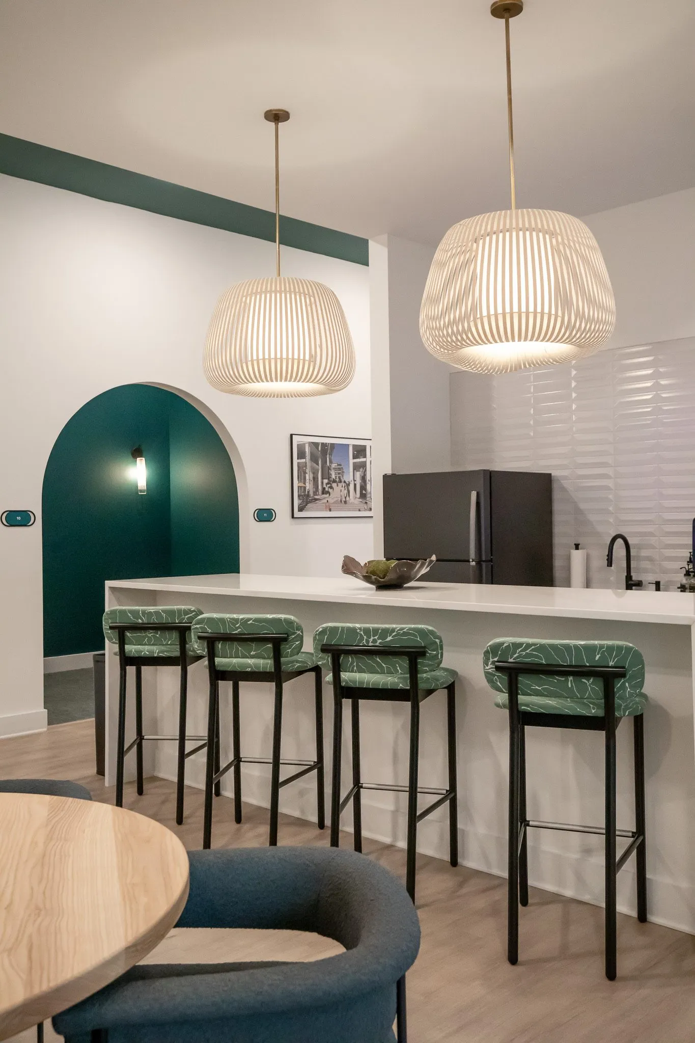 Multifamily amenity break room counter with leaf print stools under slatted pendants, a teal arch beyond