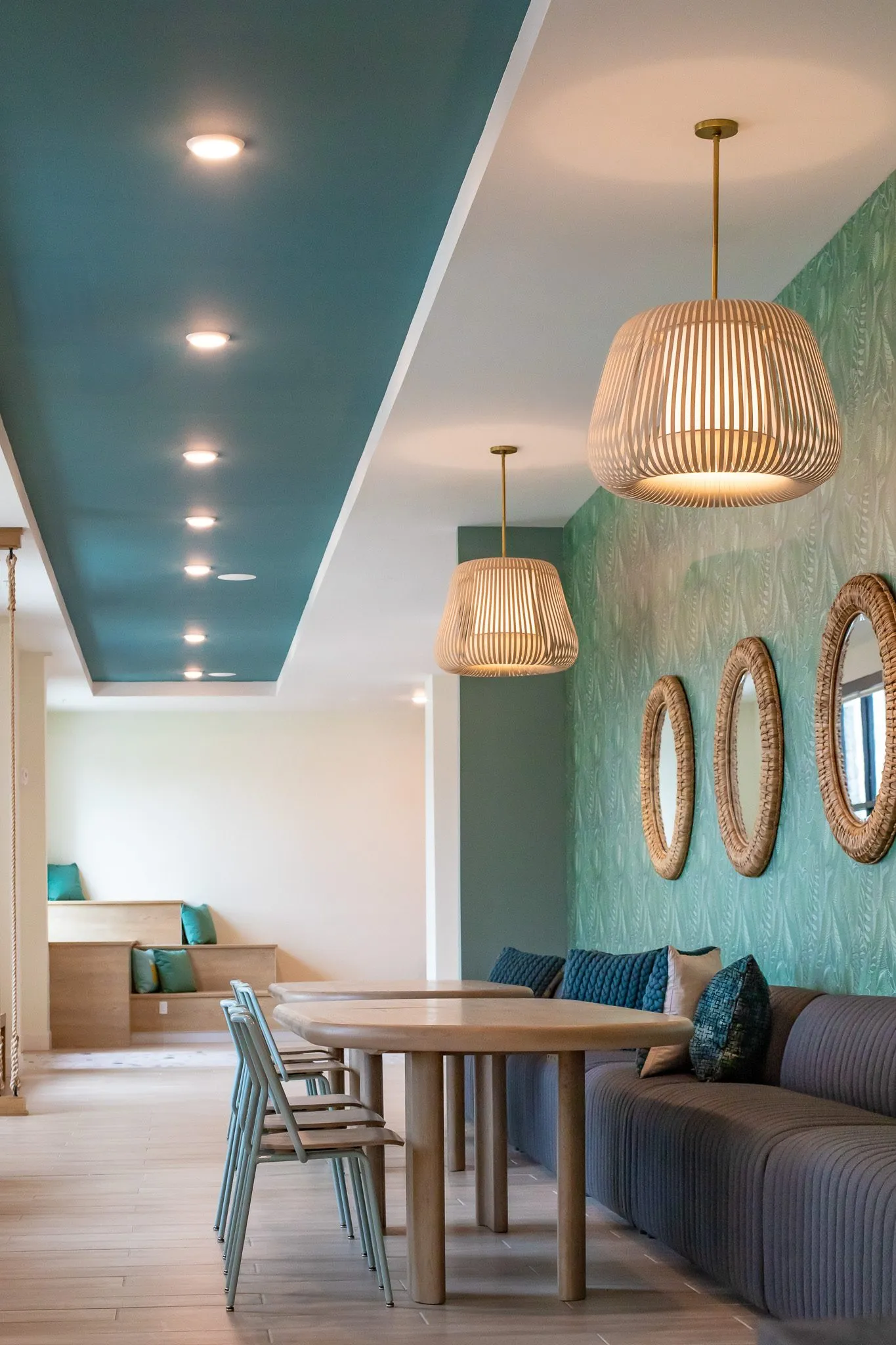 Multifamily amenity banquette hall under a teal ceiling band, slatted pendants and rattan mirrors