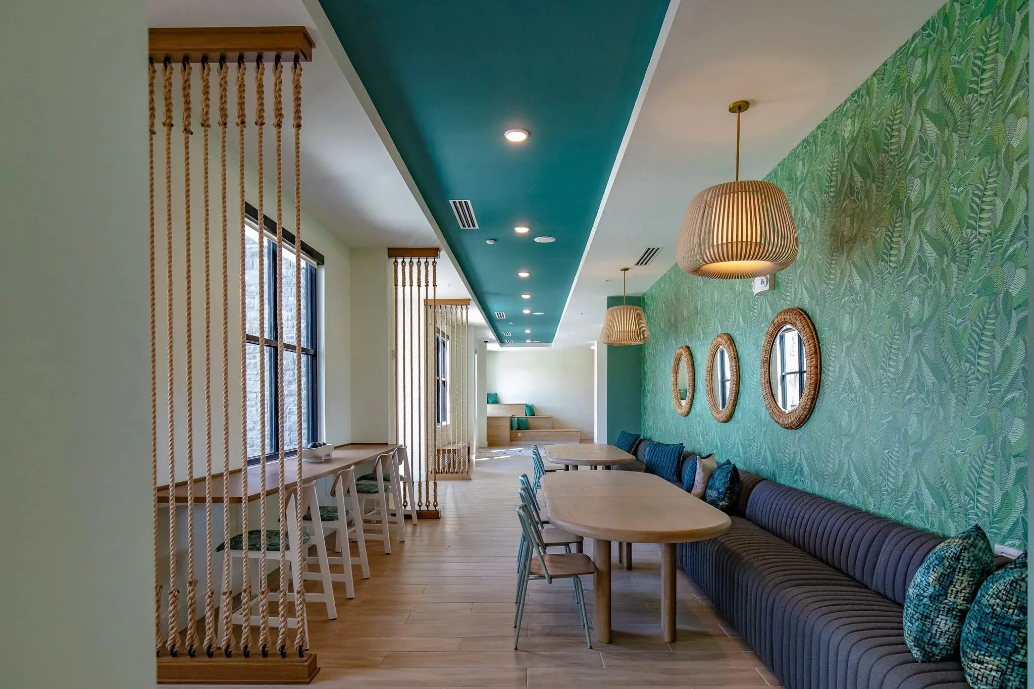 Multifamily amenity banquette hall with navy seating along a palm printed wall under woven pendants and rattan mirrors