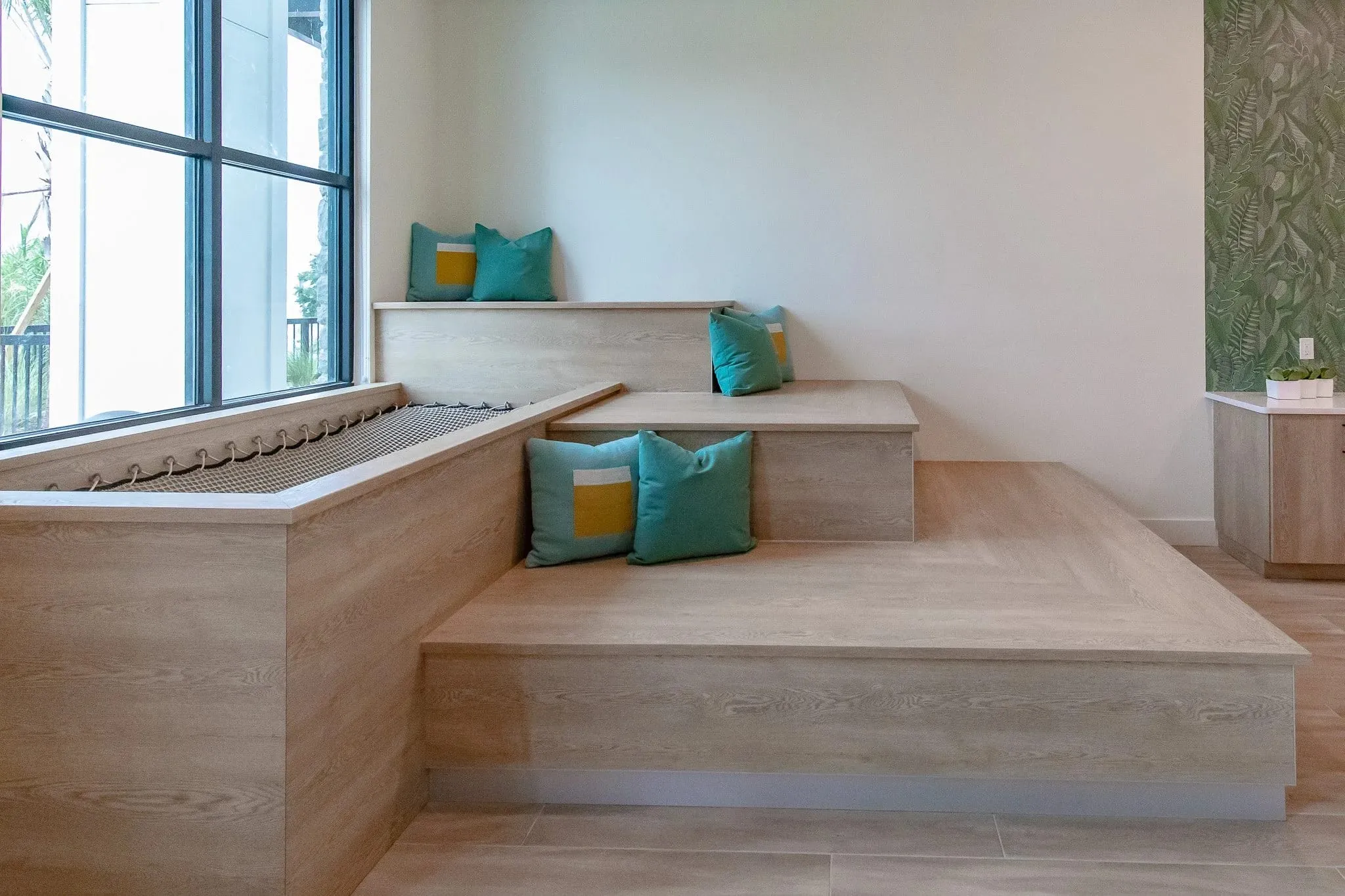 Multifamily amenity bleacher seating with pale wood steps and teal cushions beside the tall window