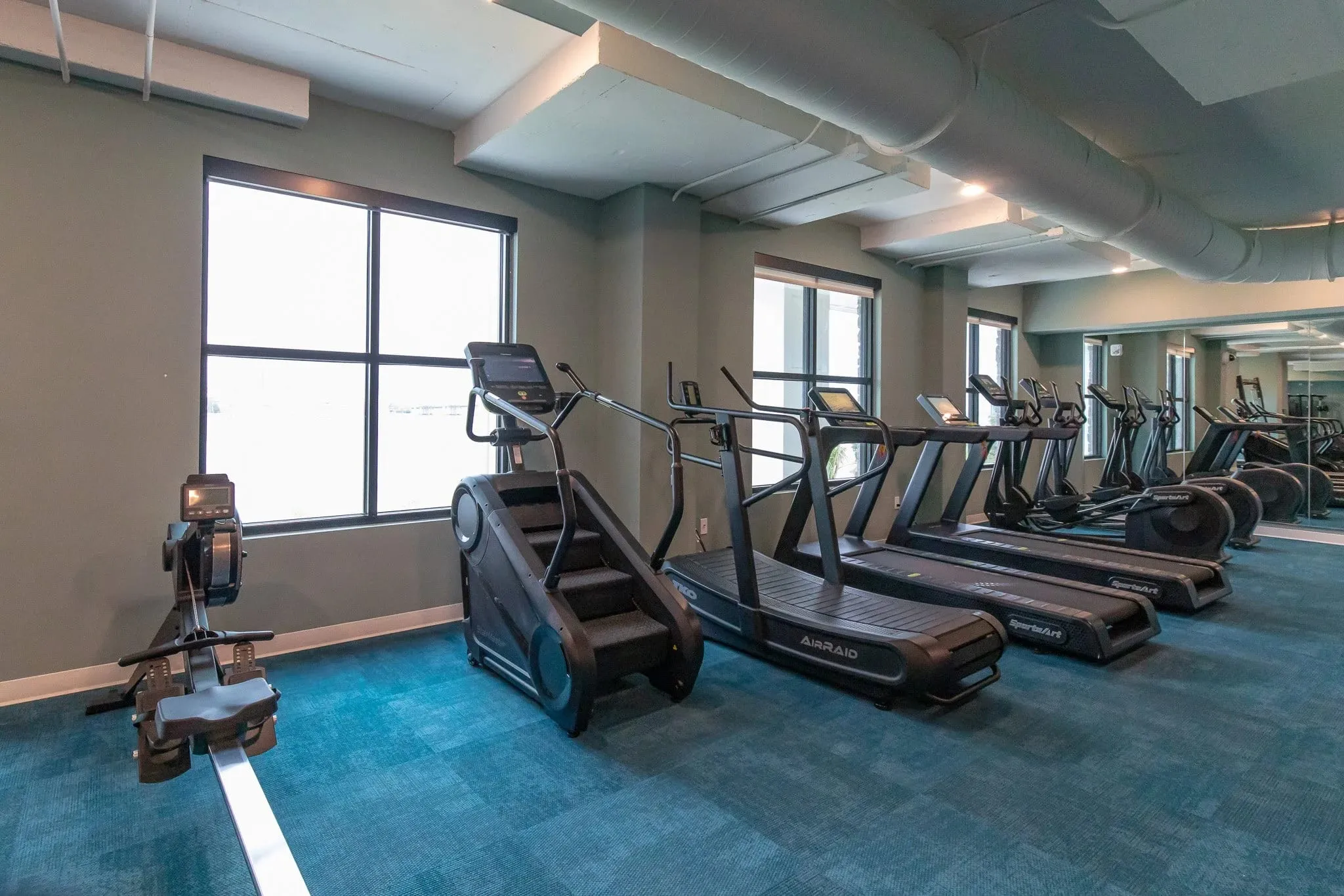 Multifamily amenity fitness center with treadmills and a stair climber along the windows above a teal floor