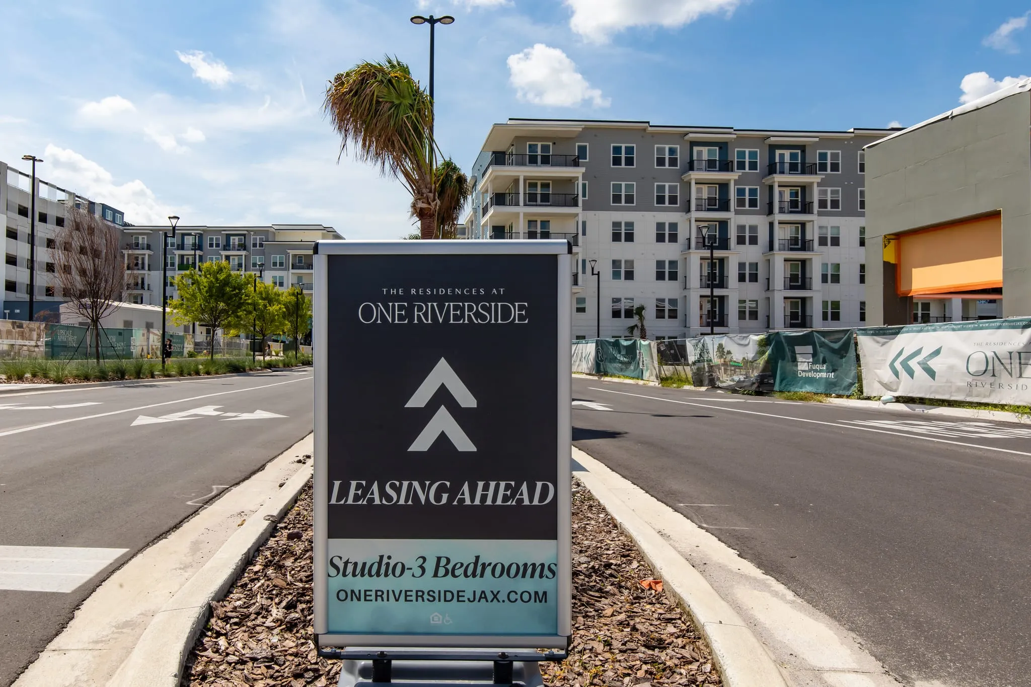 Multifamily community exterior: leasing sign with the brand identity and chevron mark on a roadside board before the building