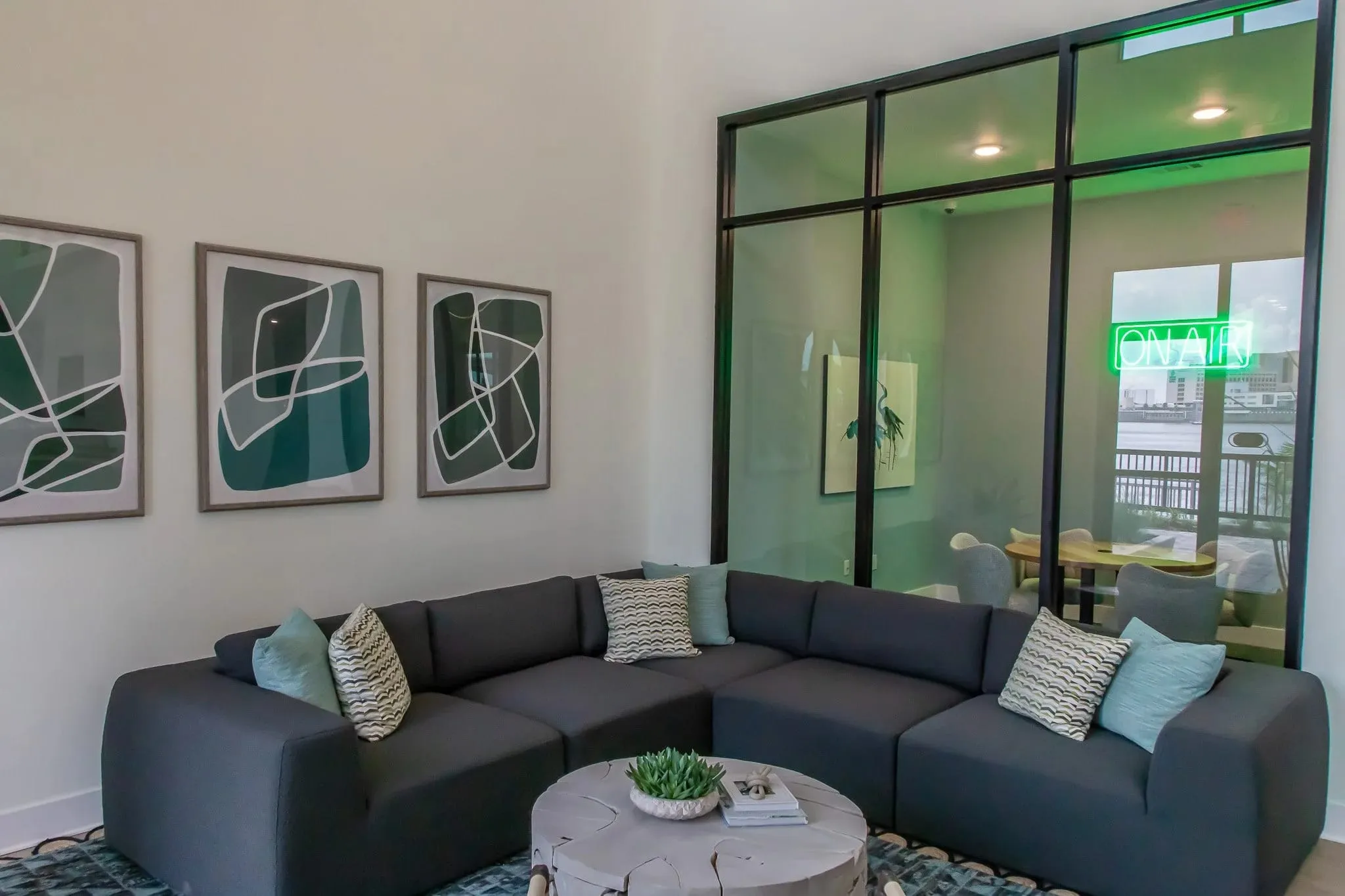 Multifamily amenity lounge with a grey sectional beside the glass-walled podcast room, its On Air neon sign glowing