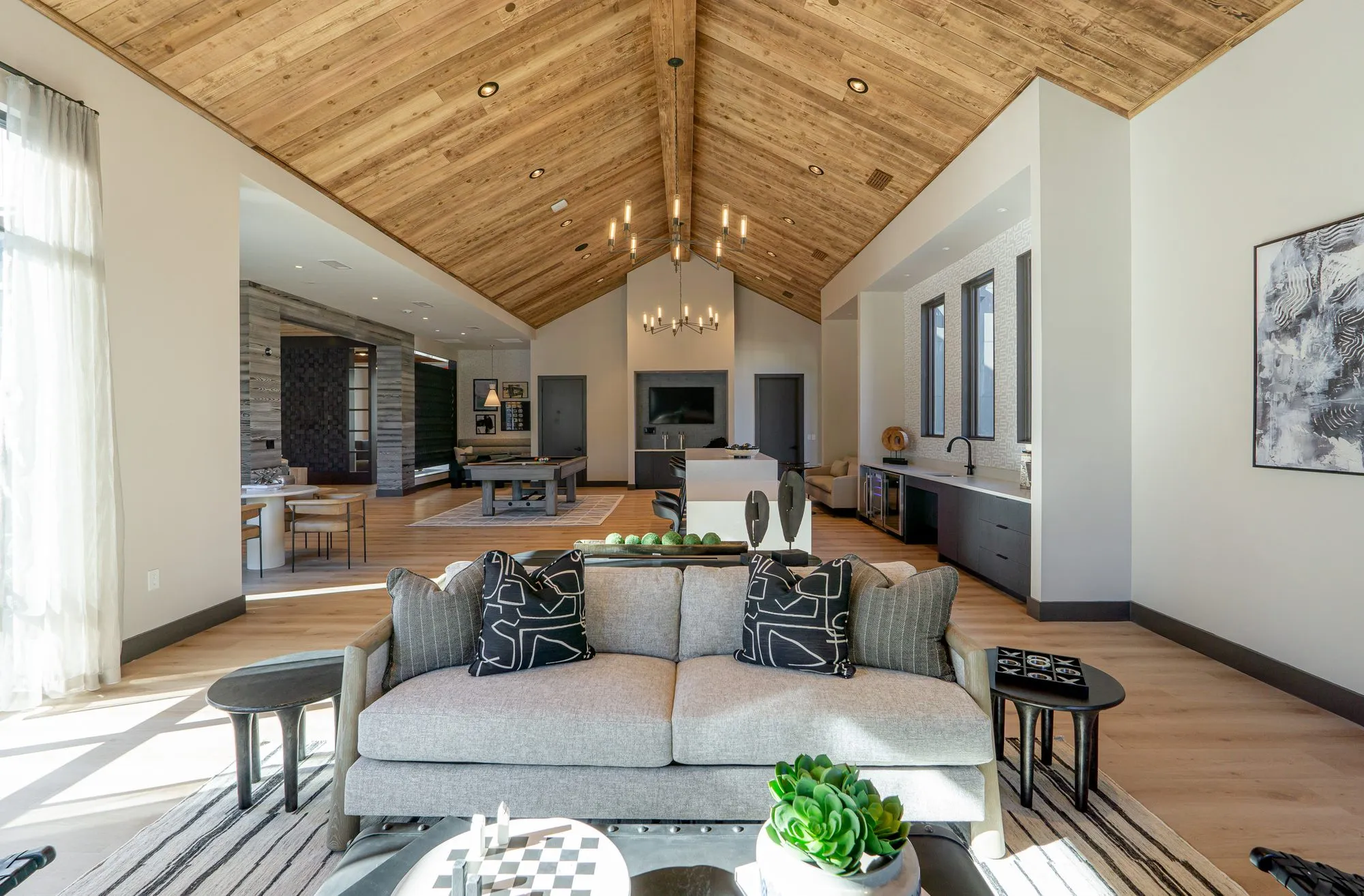 Multifamily amenity clubroom under a wood plank vaulted ceiling, grey sofa in front, pool table and fireplace beyond