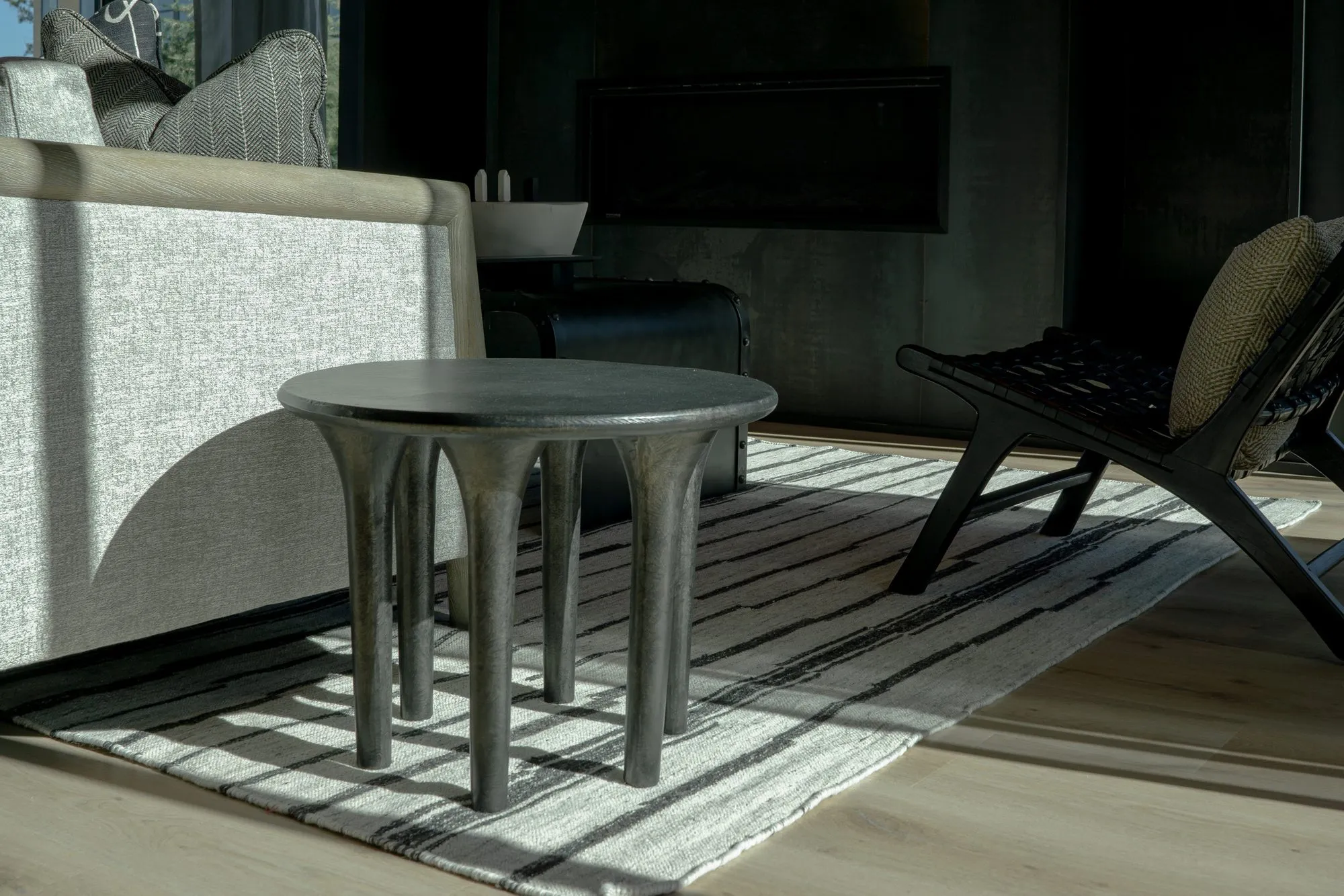 Multifamily interior detail: dark round side table with column legs on a striped rug, a sling chair and fireplace in shadow