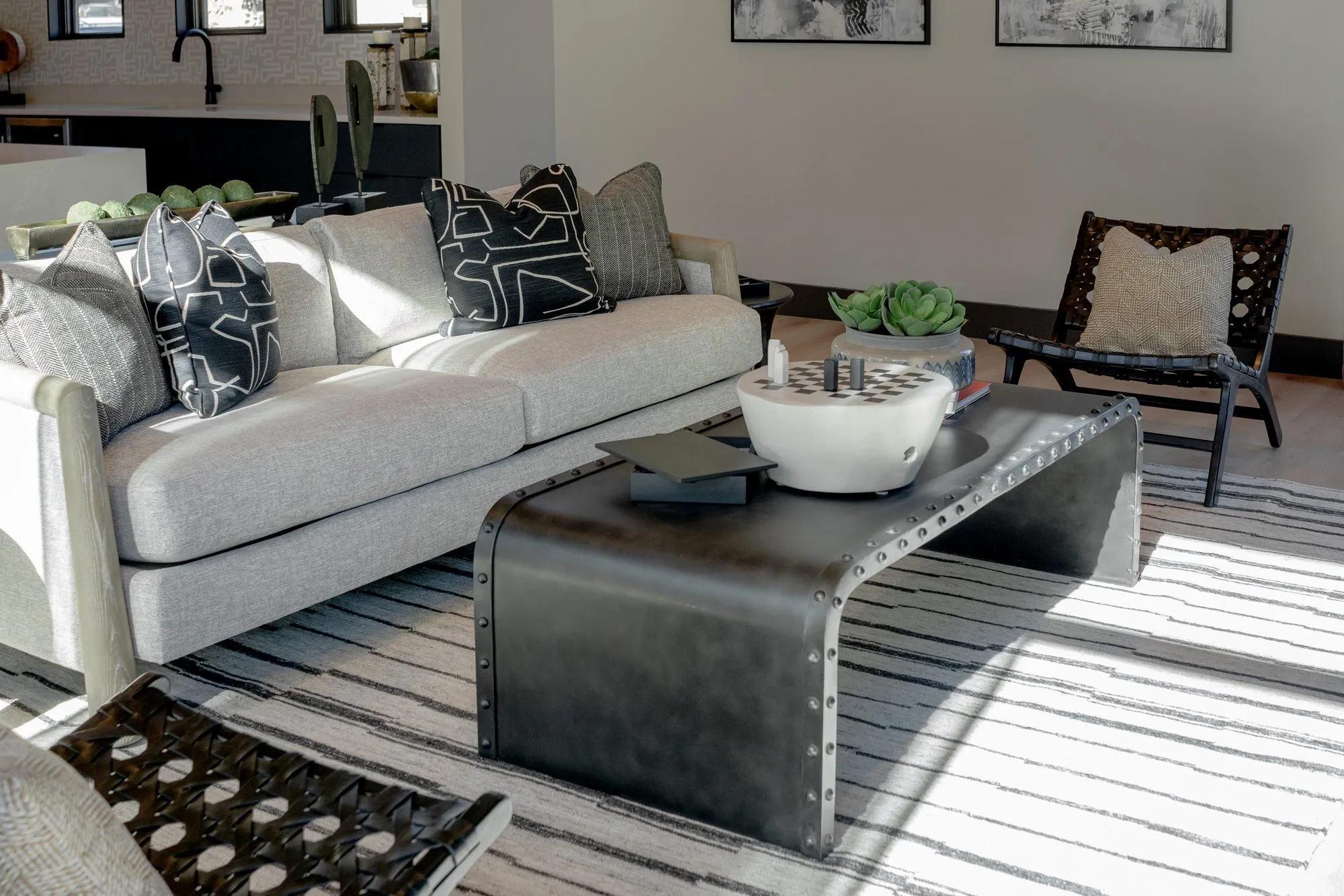 Multifamily amenity lounge with a sunlit grey sofa, a riveted black metal waterfall coffee table and a leather sling chair