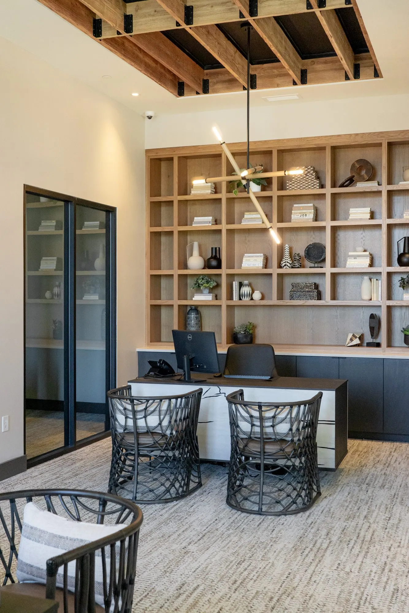 Multifamily amenity leasing office with glass doors, oak shelving and a desk with two woven chairs under timber joists