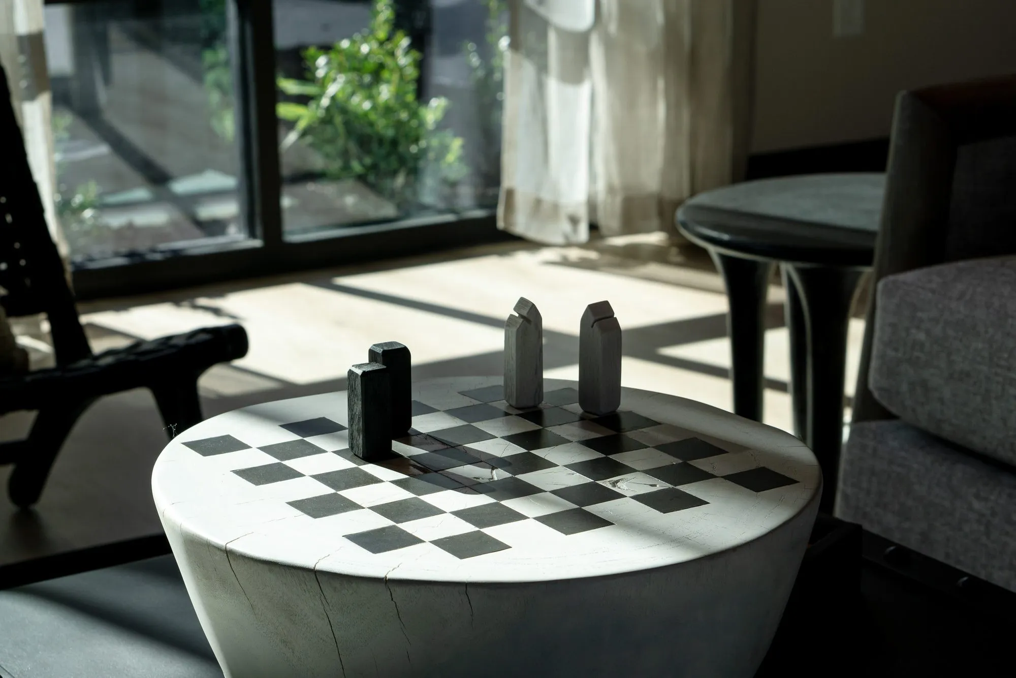 Multifamily interior detail: sunlit checkerboard drum table with game pieces beside a grey sofa, a sling chair by the window