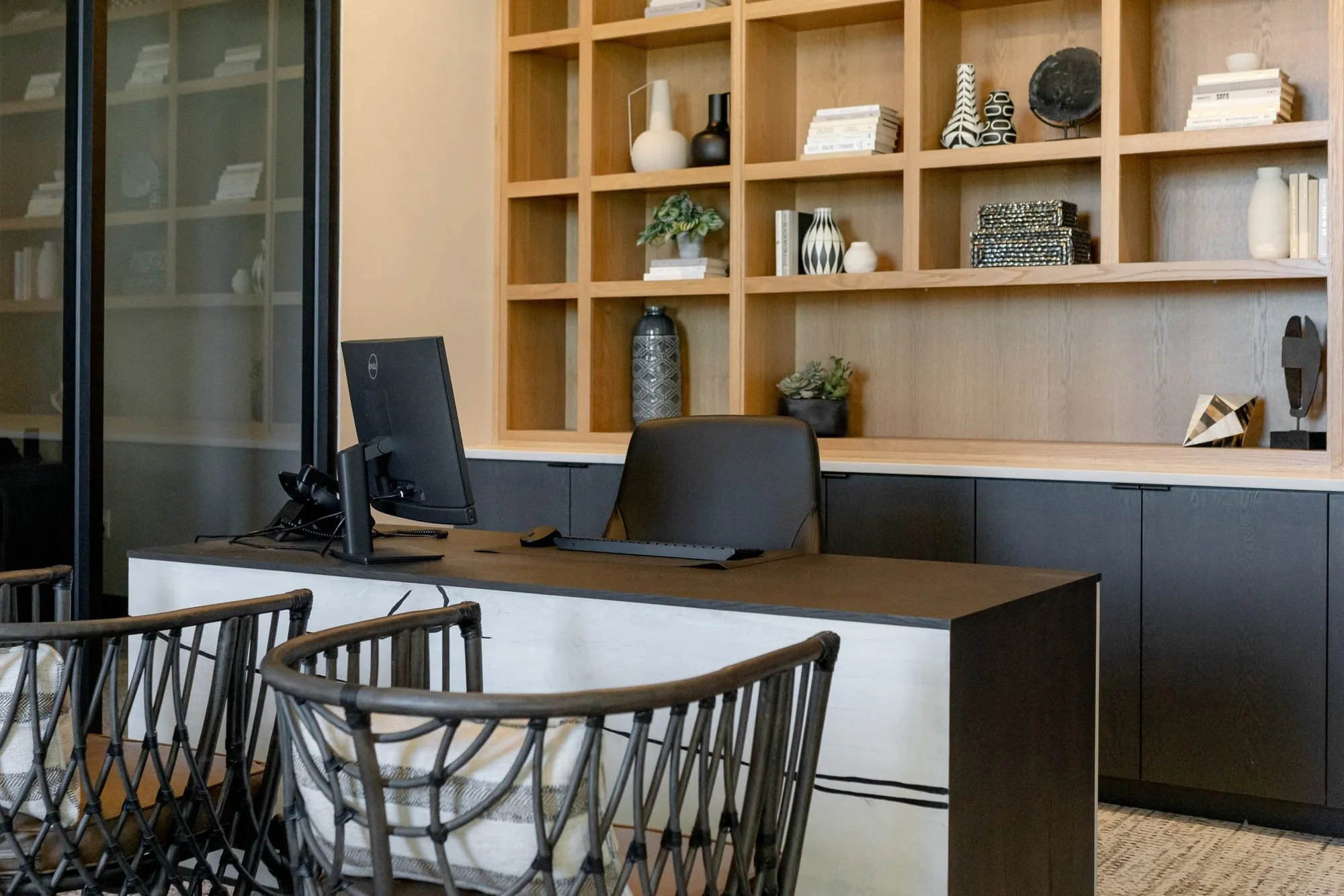 Multifamily amenity leasing desk with a monitor and two woven black chairs before oak shelving with ceramics