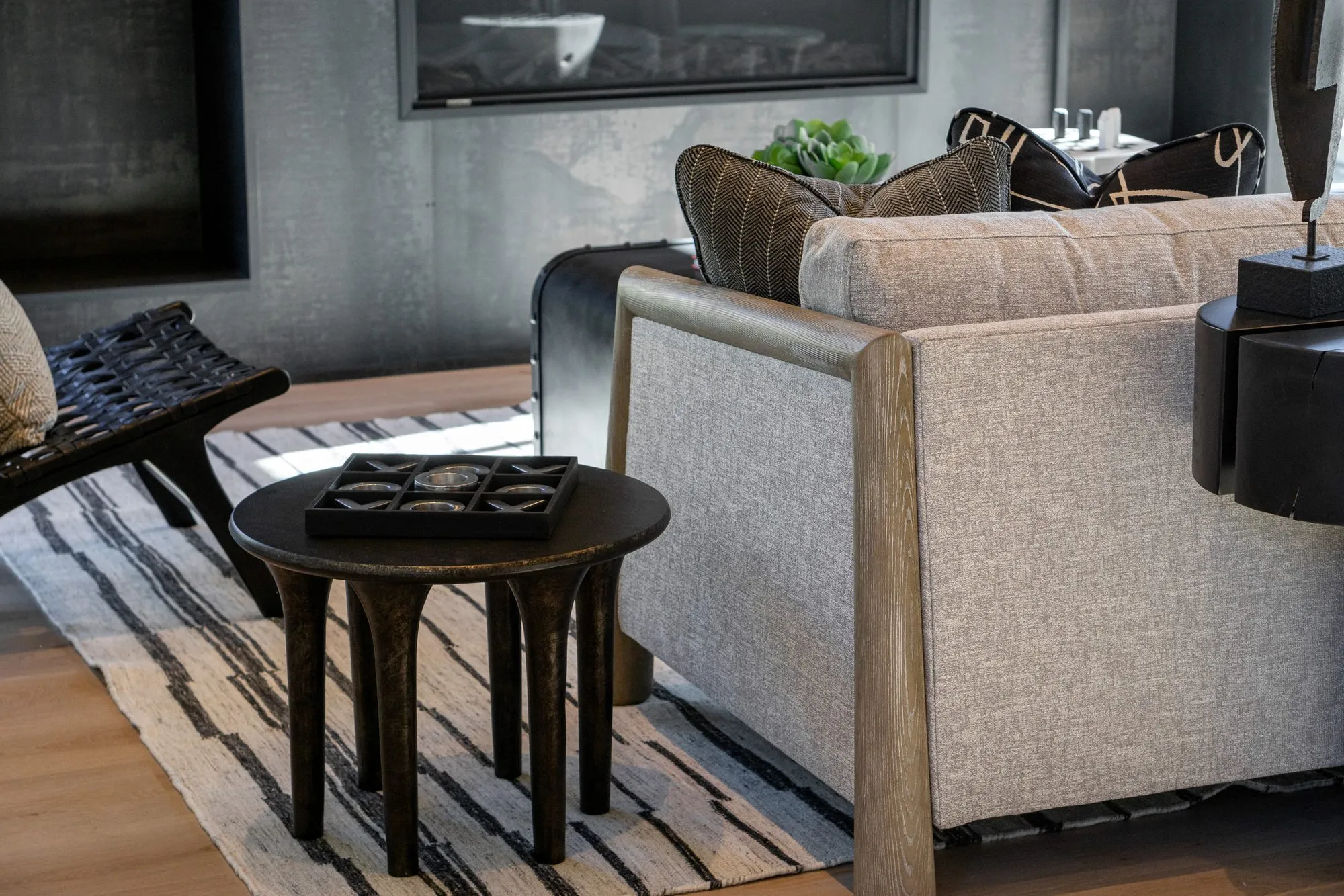 Multifamily amenity lounge with a grey wood-framed sofa and black side table on a striped rug, a sling chair and fireplace