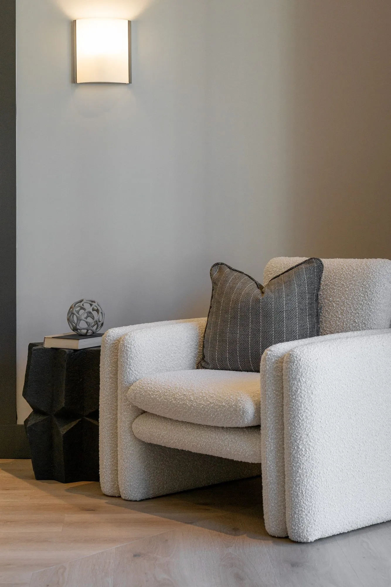 Multifamily interior detail: cream boucle armchair beside a black faceted side table below a wall sconce