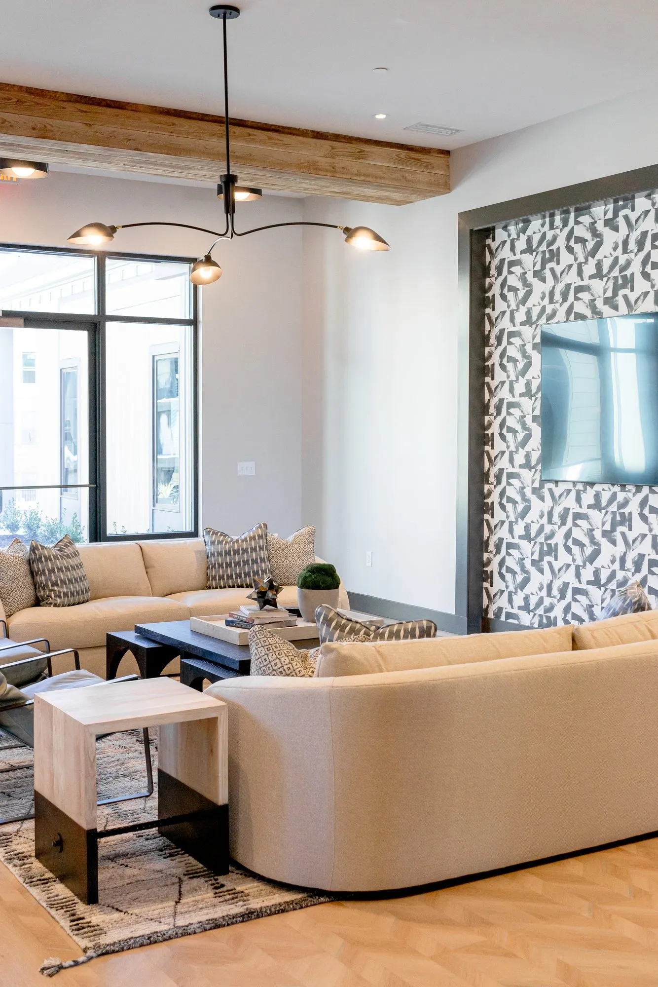 Multifamily amenity lounge with cream sofas under a multi-arm pendant and timber beam, TV on a patterned tile wall