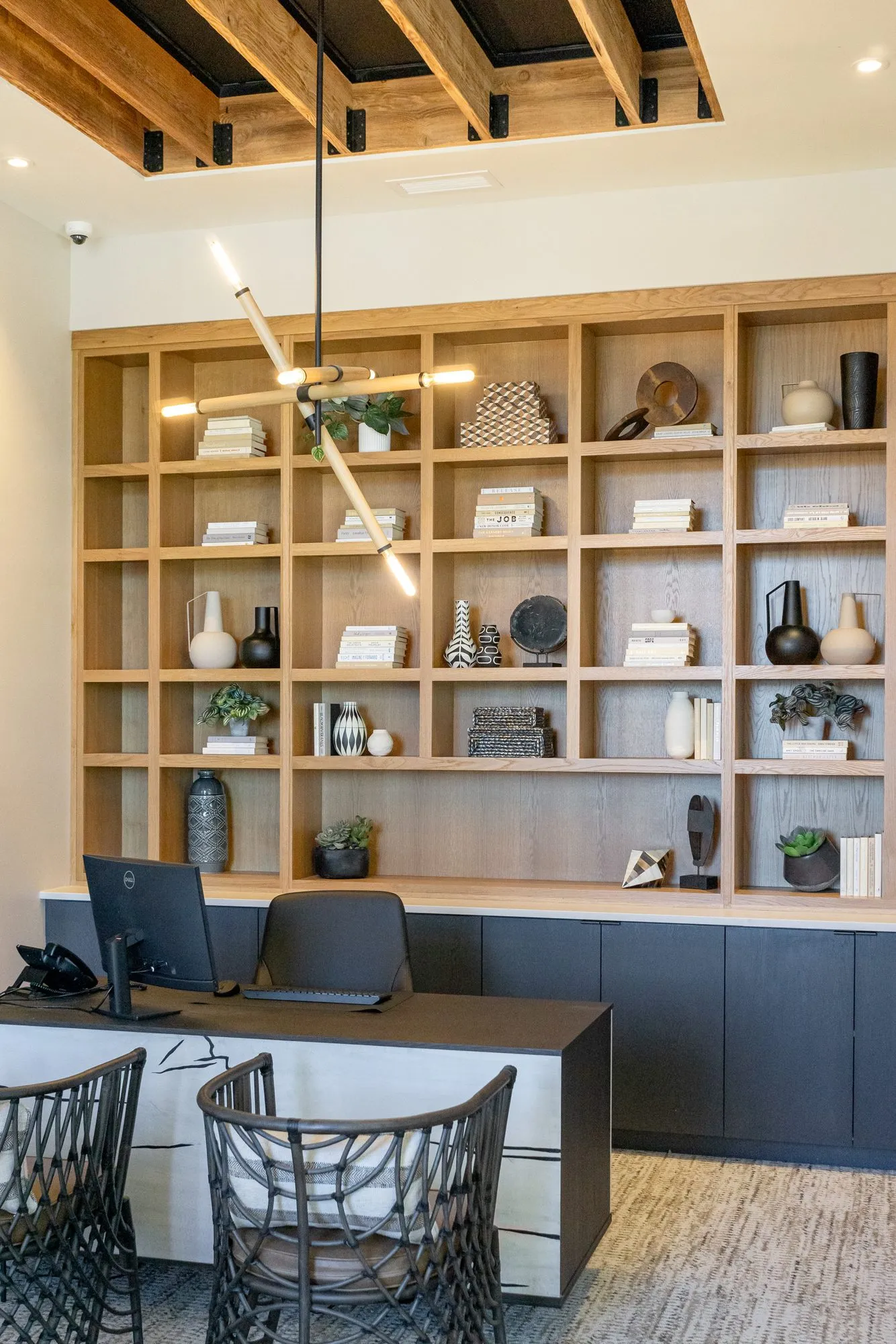 Multifamily amenity office with an oak shelving wall above dark cabinets, a desk and woven chairs, a linear pendant in front