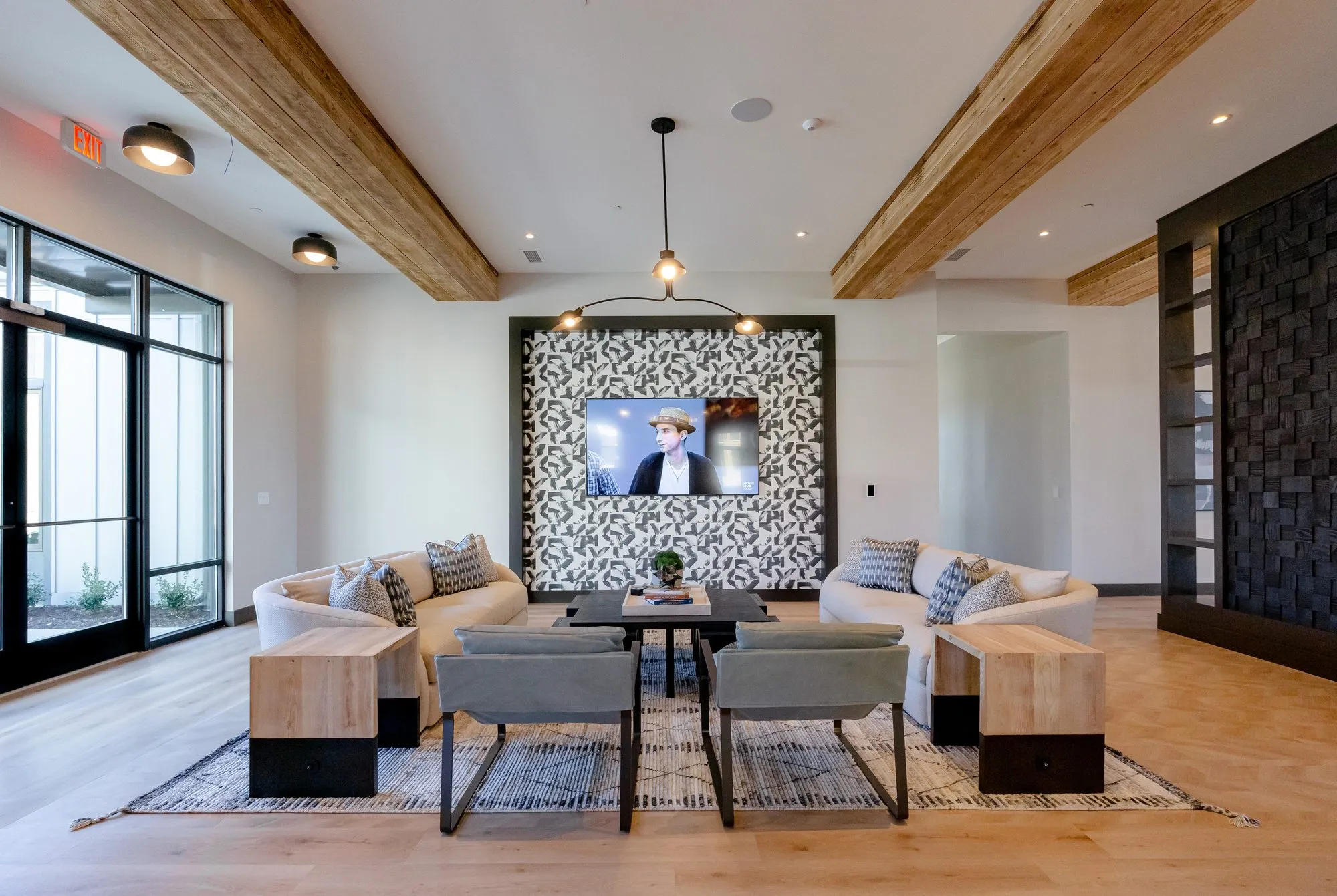 Multifamily amenity lounge with cream sofas and grey sling chairs facing a TV on a patterned tile wall, timber beams above