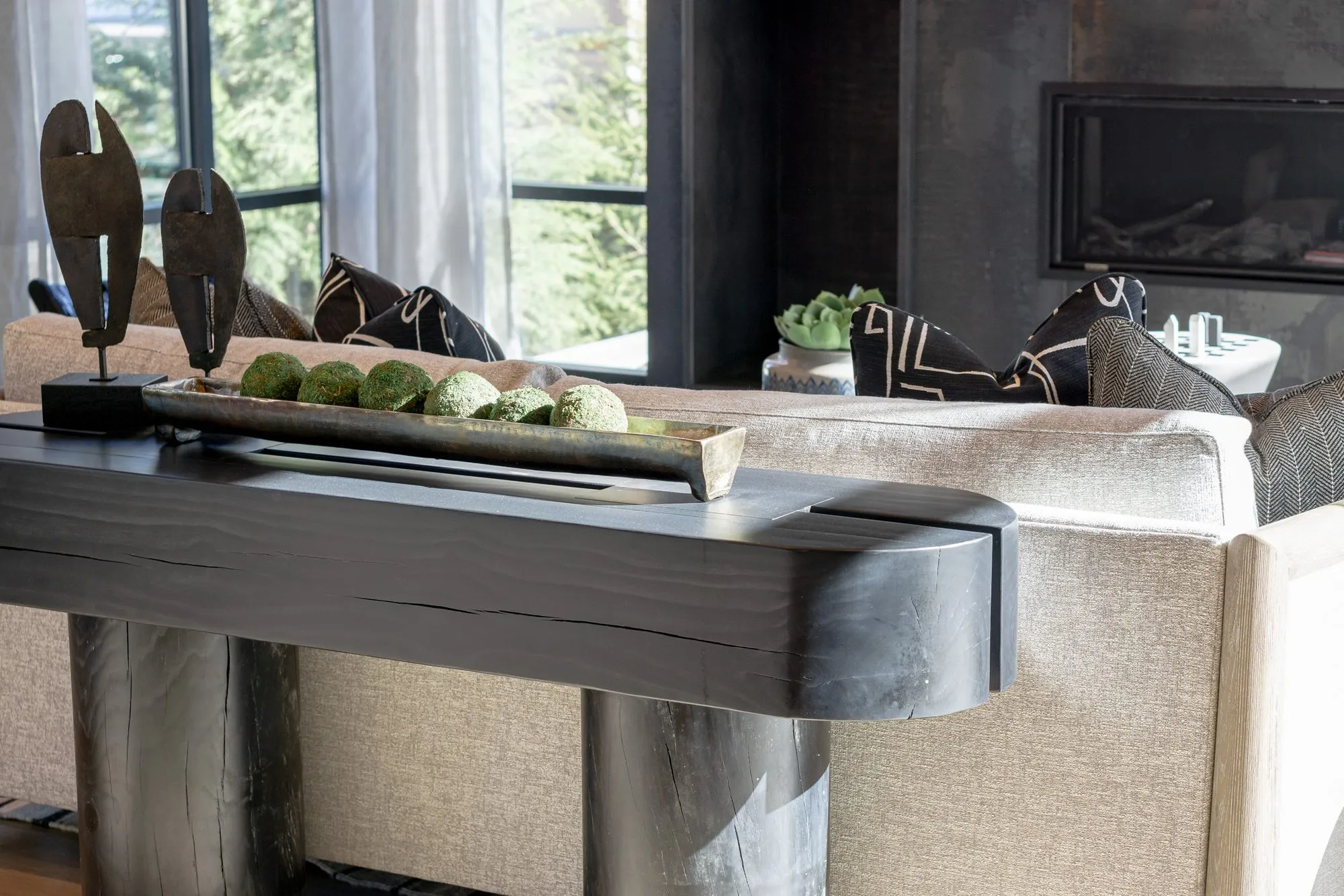 Multifamily interior detail: dark wood console with sculptures and moss balls behind a cream sofa, linear fireplace beyond